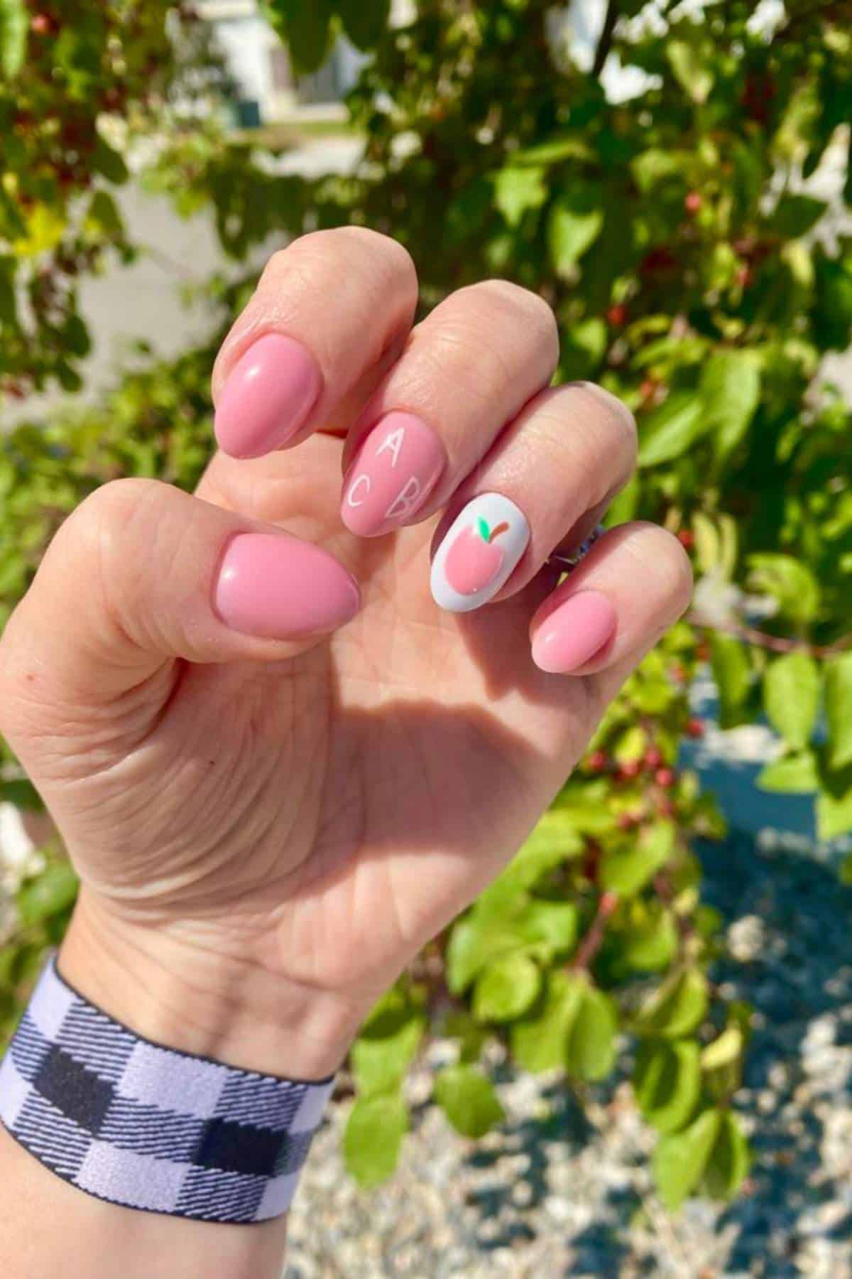 A hand with short, rounded pink nails; one nail has a peach design and the letters "A" and "B" on two other nails. A black and white plaid wristband is visible. Green foliage in background.