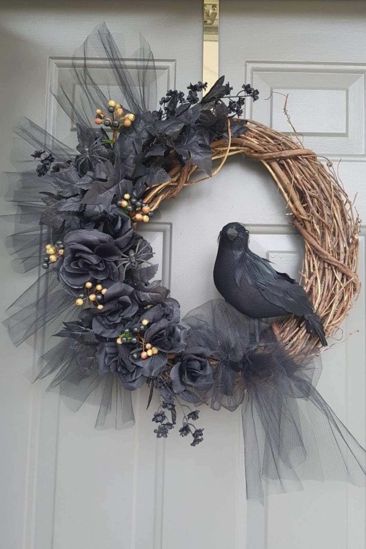 A grapevine wreath with black flowers, tulle, faux berries, and a black crow decoration hangs on a light-colored door.