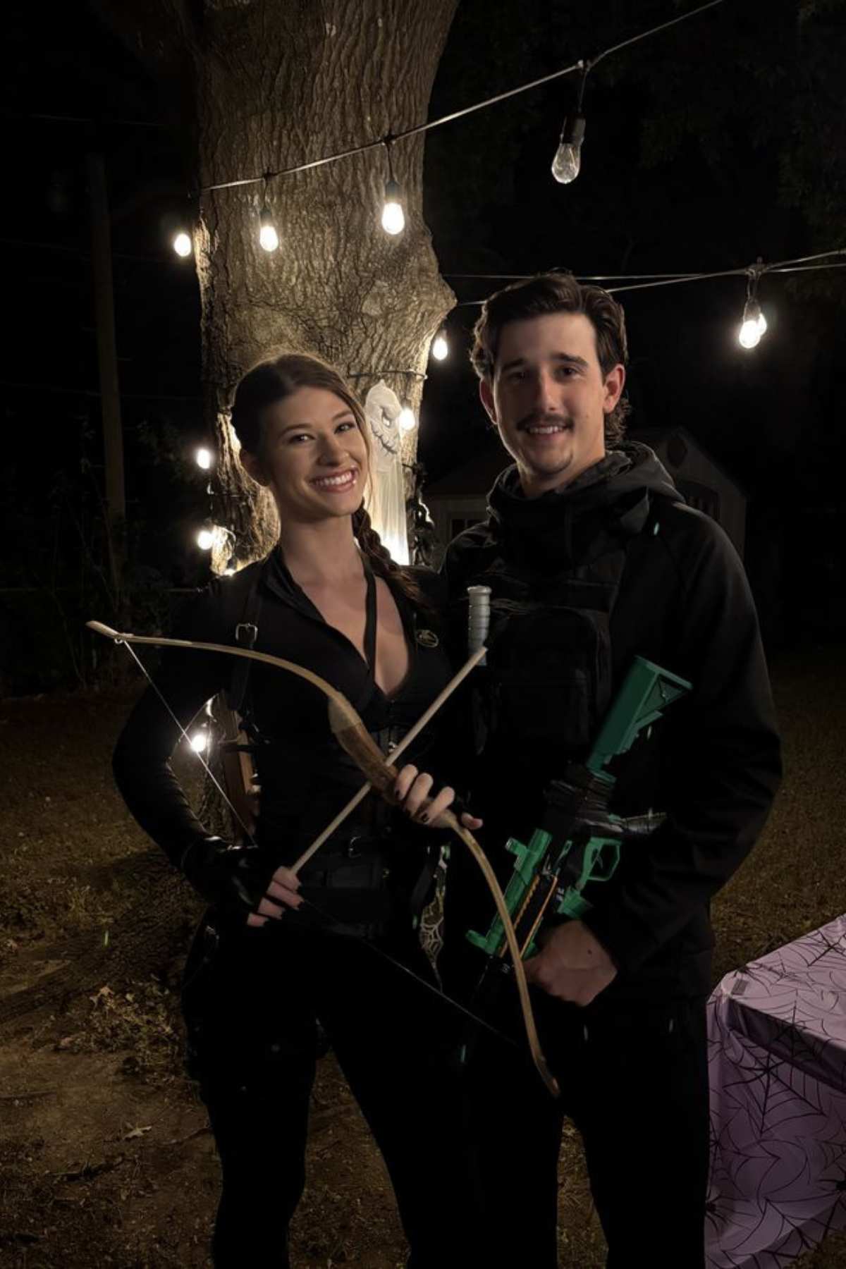 Two people in dark clothing stand outdoors at night, holding a bow and a toy gun, with string lights and a tree in the background.