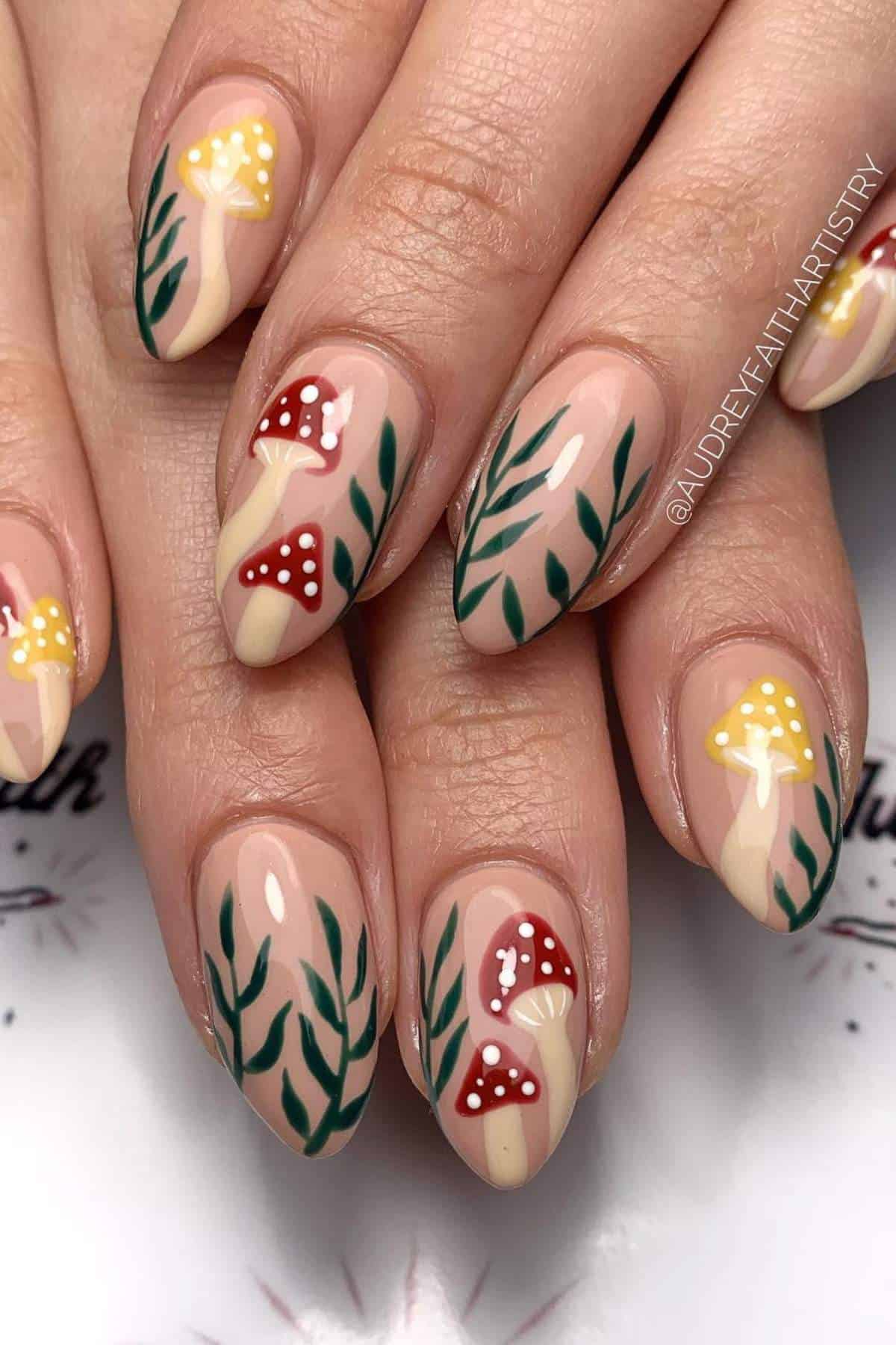 Fingernails with nude polish feature painted red and yellow mushrooms with white spots and green leaves.