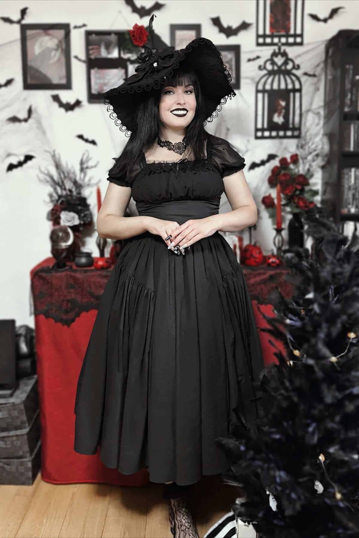 Woman in a black Victorian-style dress and hat stands indoors, surrounded by gothic Halloween decorations including bats, candles, and black-and-red flowers.