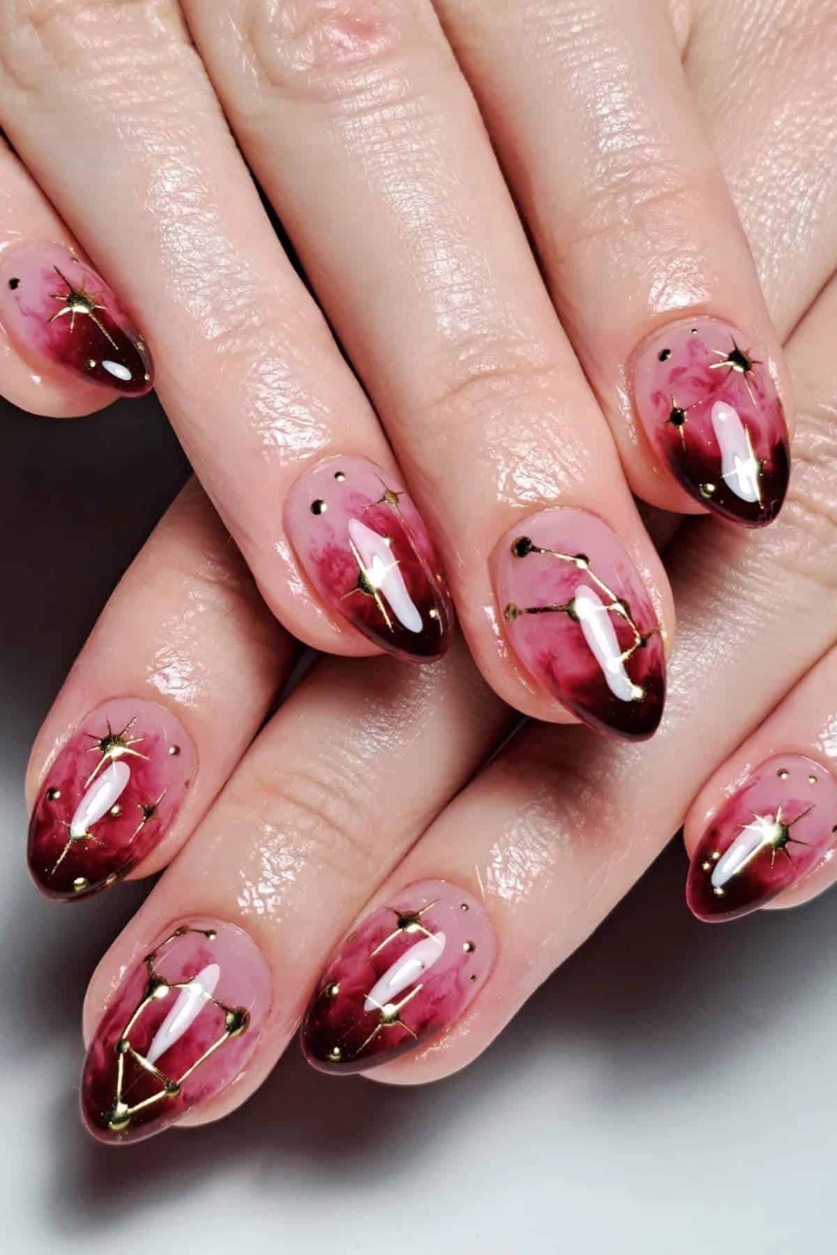 Hands with almond-shaped nails painted in pink and maroon ombre, featuring gold star and constellation designs on each nail.