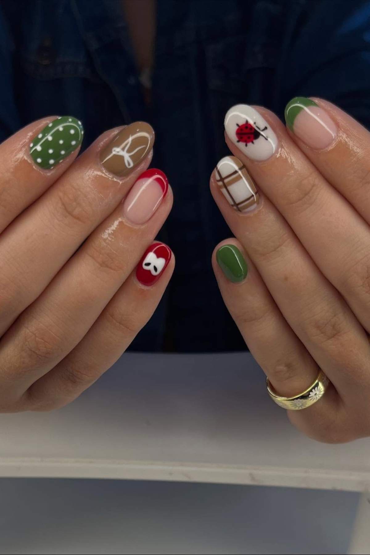 Close-up of hands with manicured nails featuring various designs, including green with white dots, a white bow, a ladybug, a red apple, and a brown plaid pattern.