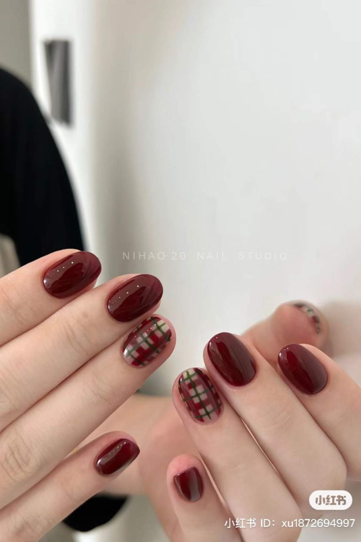 Hands with glossy dark red manicure; ring fingers feature a plaid design in red, green, and white.