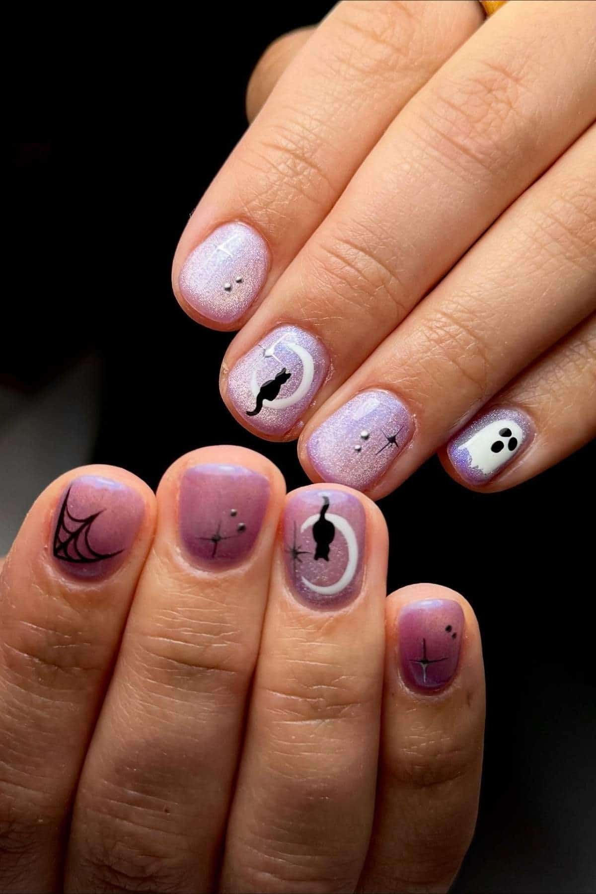 Close-up of hands with short nails painted in shimmery lavender polish, featuring Halloween designs like a ghost, spiderweb, bat, cat, and abstract art on each nail.