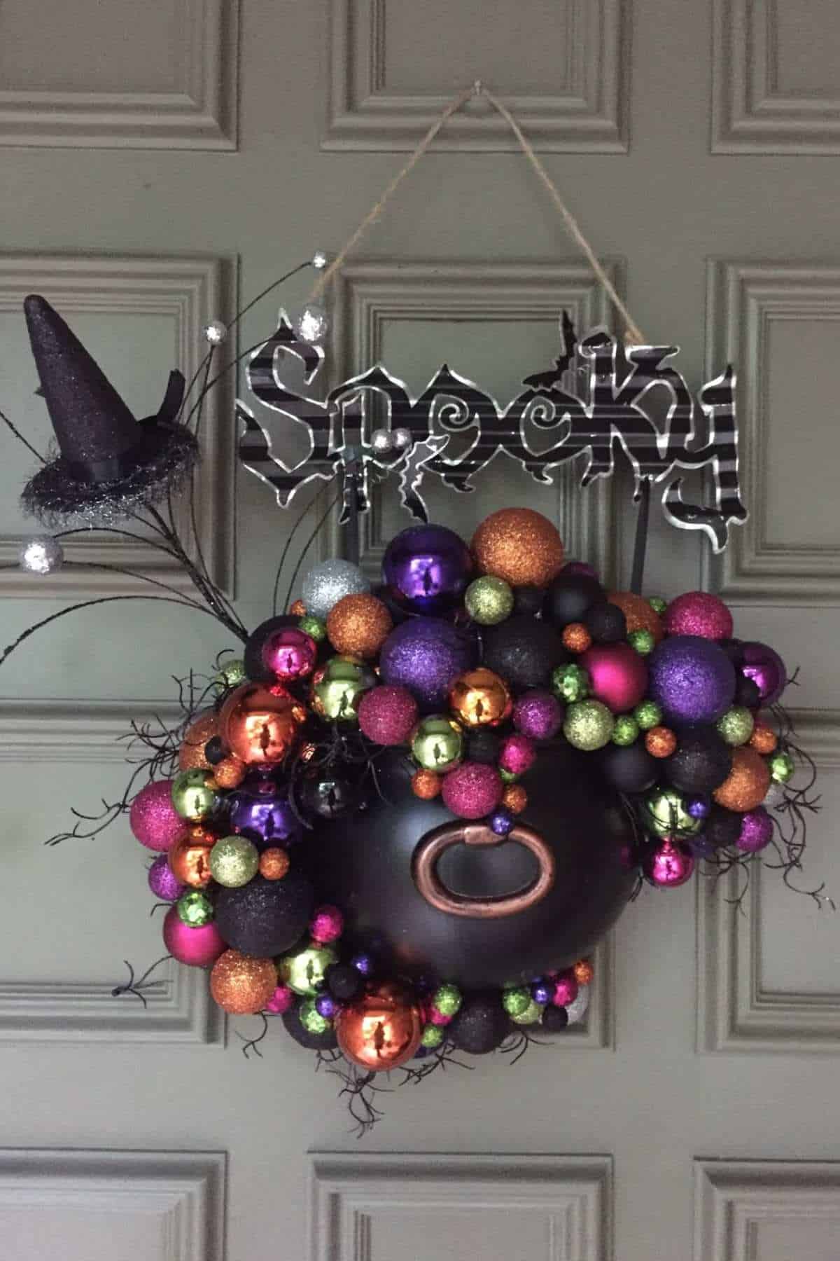 A Halloween wreath made of colorful ornaments with a black cauldron center, a small witch hat, and a hanging sign that reads “Spooky” on a paneled door.