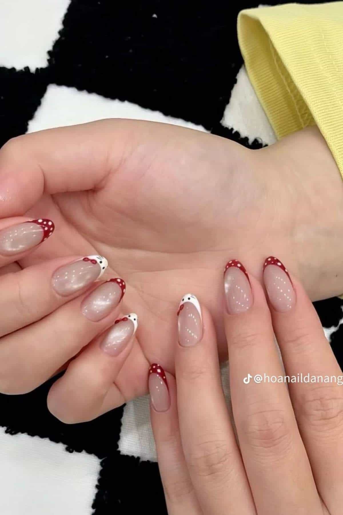 Close-up of hands with almond-shaped nails featuring red and white French tips, some with polka dots, on a black and white checkered surface.