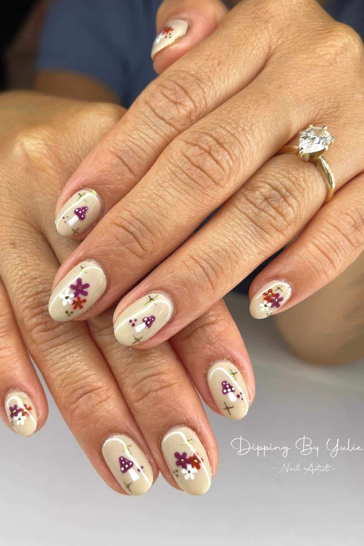 Hands with almond-shaped nails painted a light beige polish, each nail featuring small purple and red floral designs. A gold ring with a large clear stone is worn on one finger.