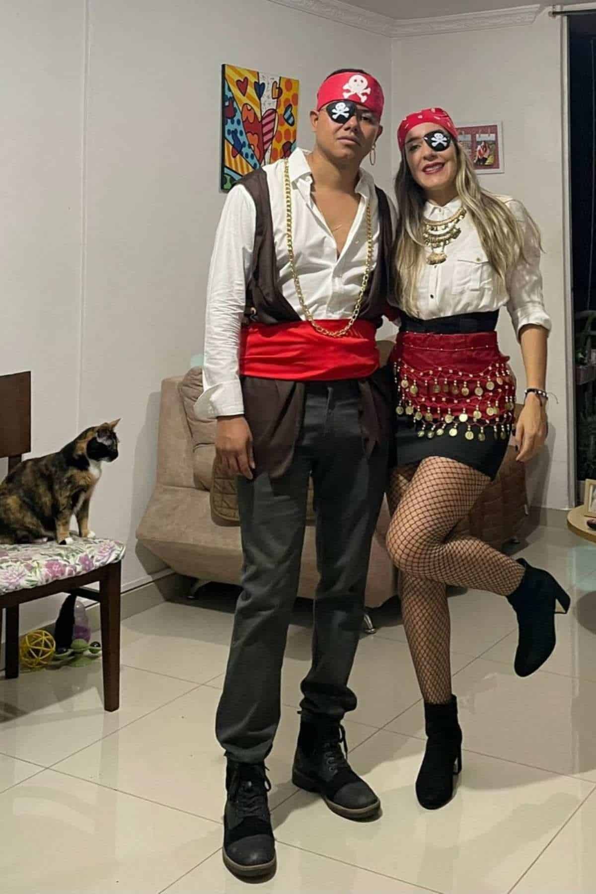 Two people dressed as pirates pose indoors, wearing eye patches and bandanas. A cat sits on a chair nearby.