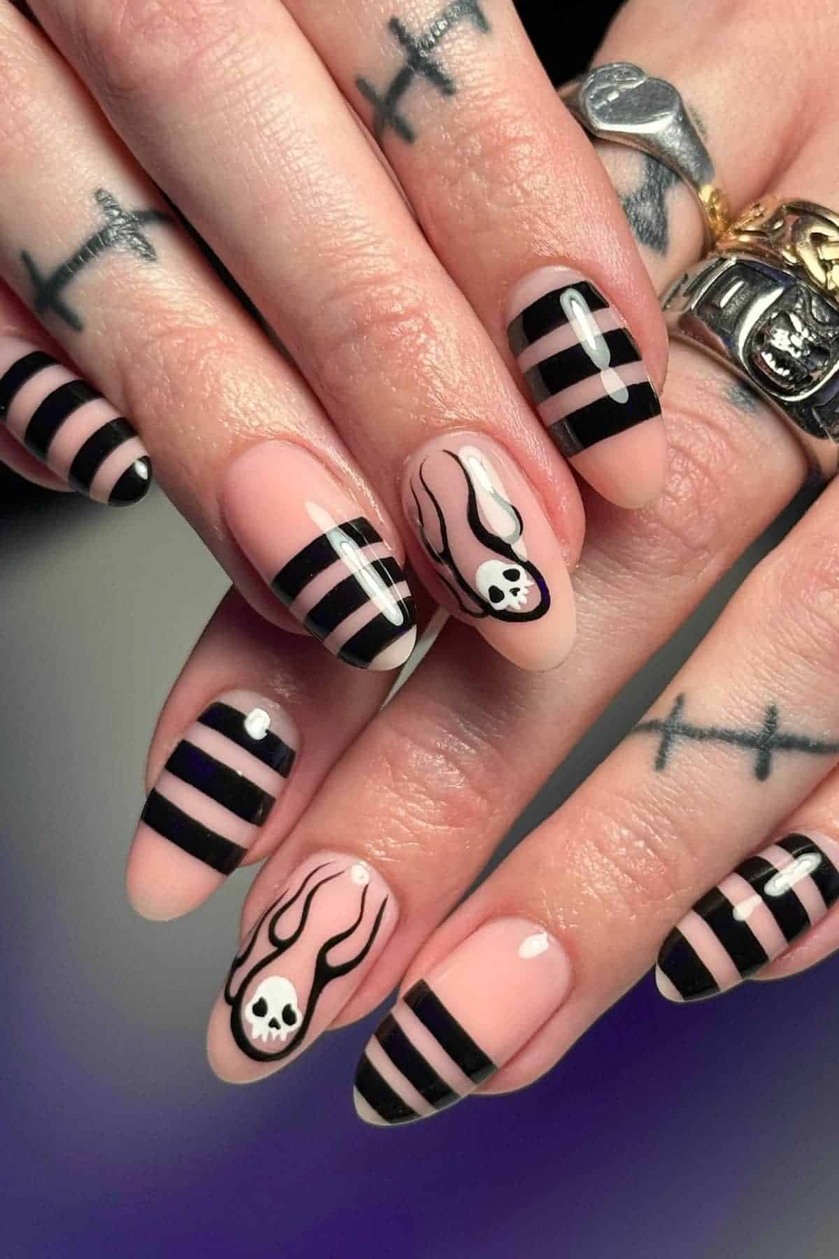 Hands with long, almond-shaped nails painted with black stripes and skull designs. Fingers have tattooed crosses and wear silver rings.