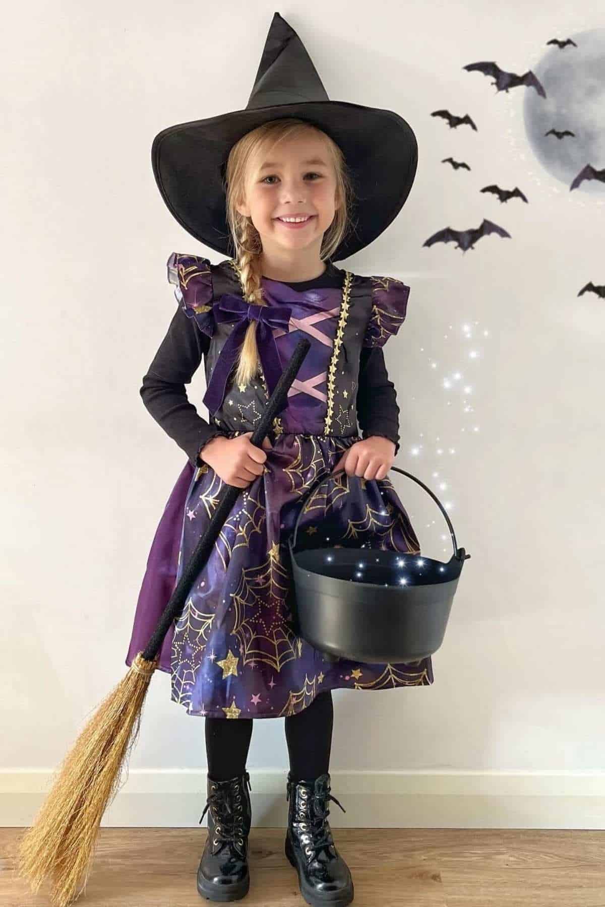 Young girl dressed as a witch with a black hat, purple spiderweb dress, holding a broom and black bucket, standing in front of a wall with bat decorations.