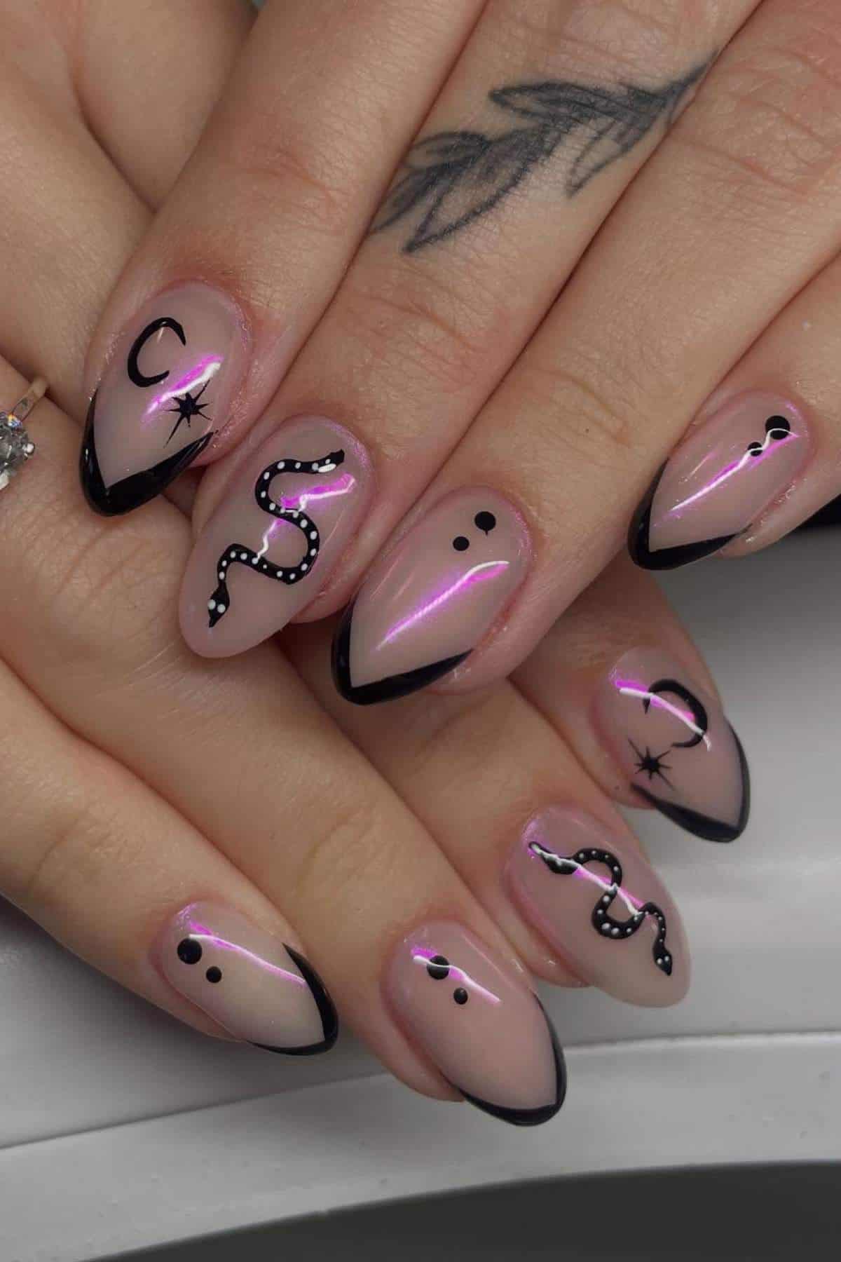Hands with almond-shaped nails, featuring black French tips, snake and dot designs, crescent moons, and stars on a shimmery nude base. A tattoo is visible on one finger.