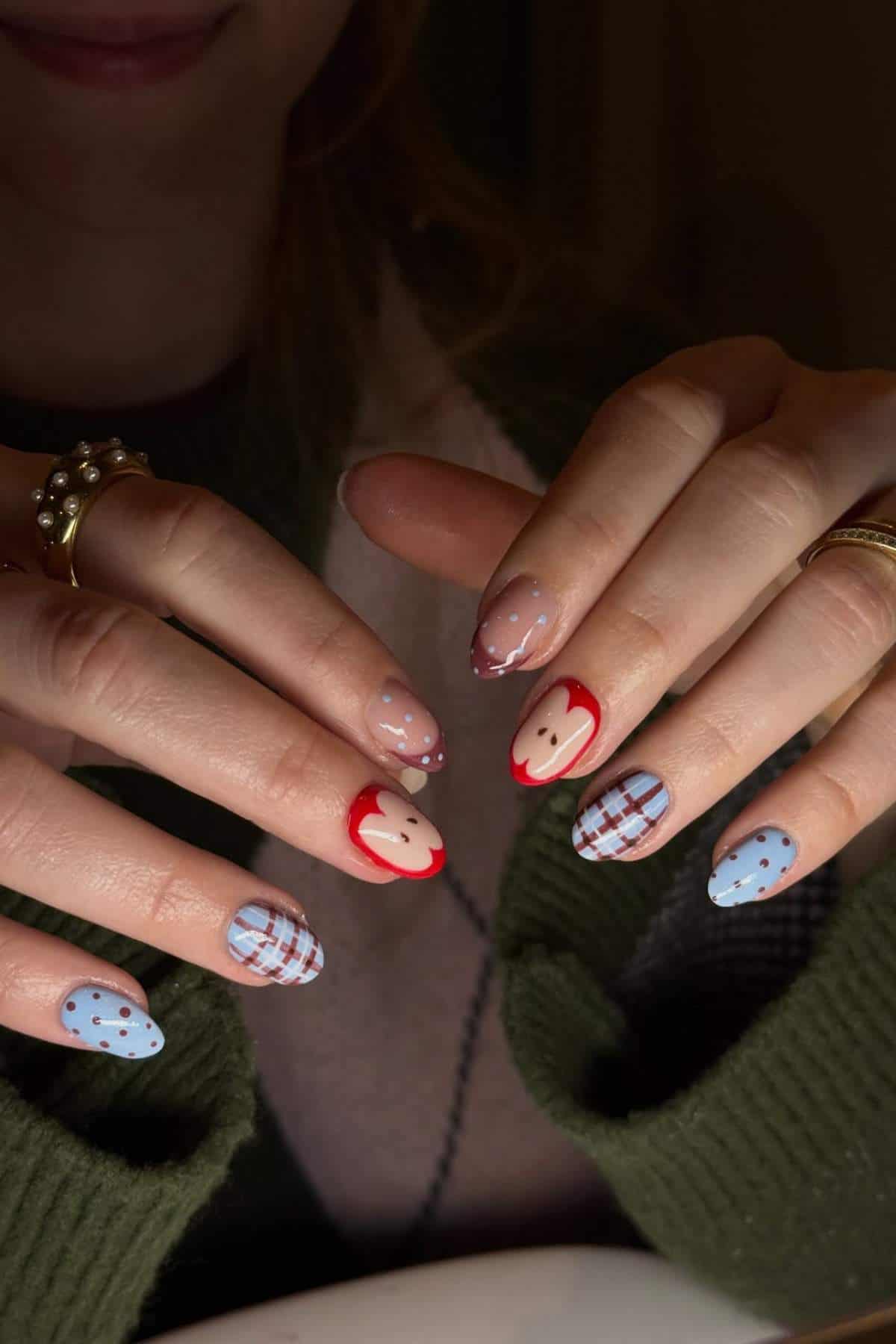 A person displays manicured nails with various designs, including red apple tips, blue with polka dots, and blue plaid patterns.