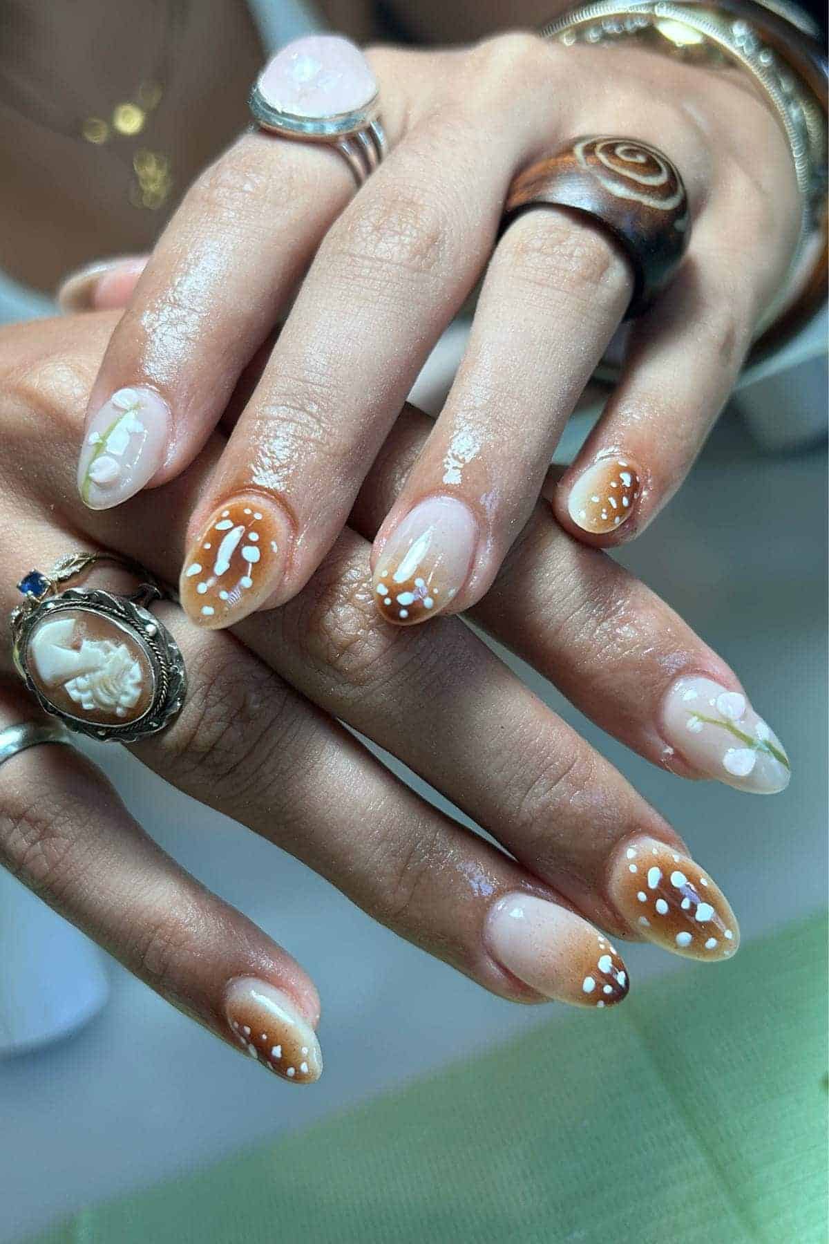 Close-up of hands with almond-shaped nails featuring brown, beige, and white abstract and dotted nail art; person is wearing multiple rings.