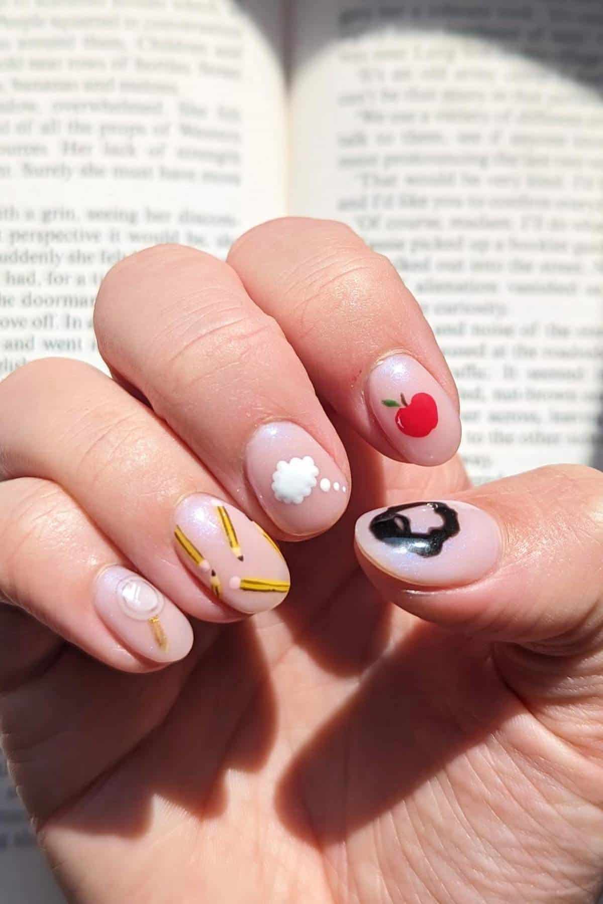 A hand with manicured nails featuring playful designs: a cloud, apple, frying pan with egg, sunny-side up egg, and French fries, held over an open book in sunlight.