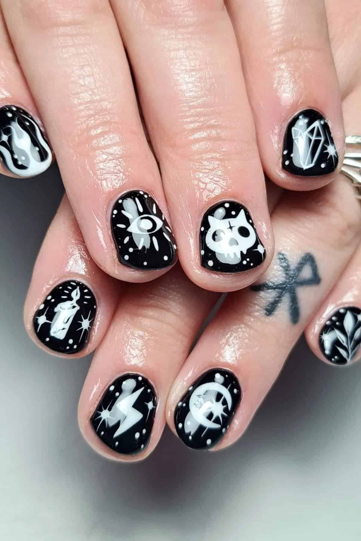 Black nails with white mystical-themed designs, including an eye, skull, diamond, lightning bolt, crescent moon, and stars. The person has a crossed lines tattoo on one finger.