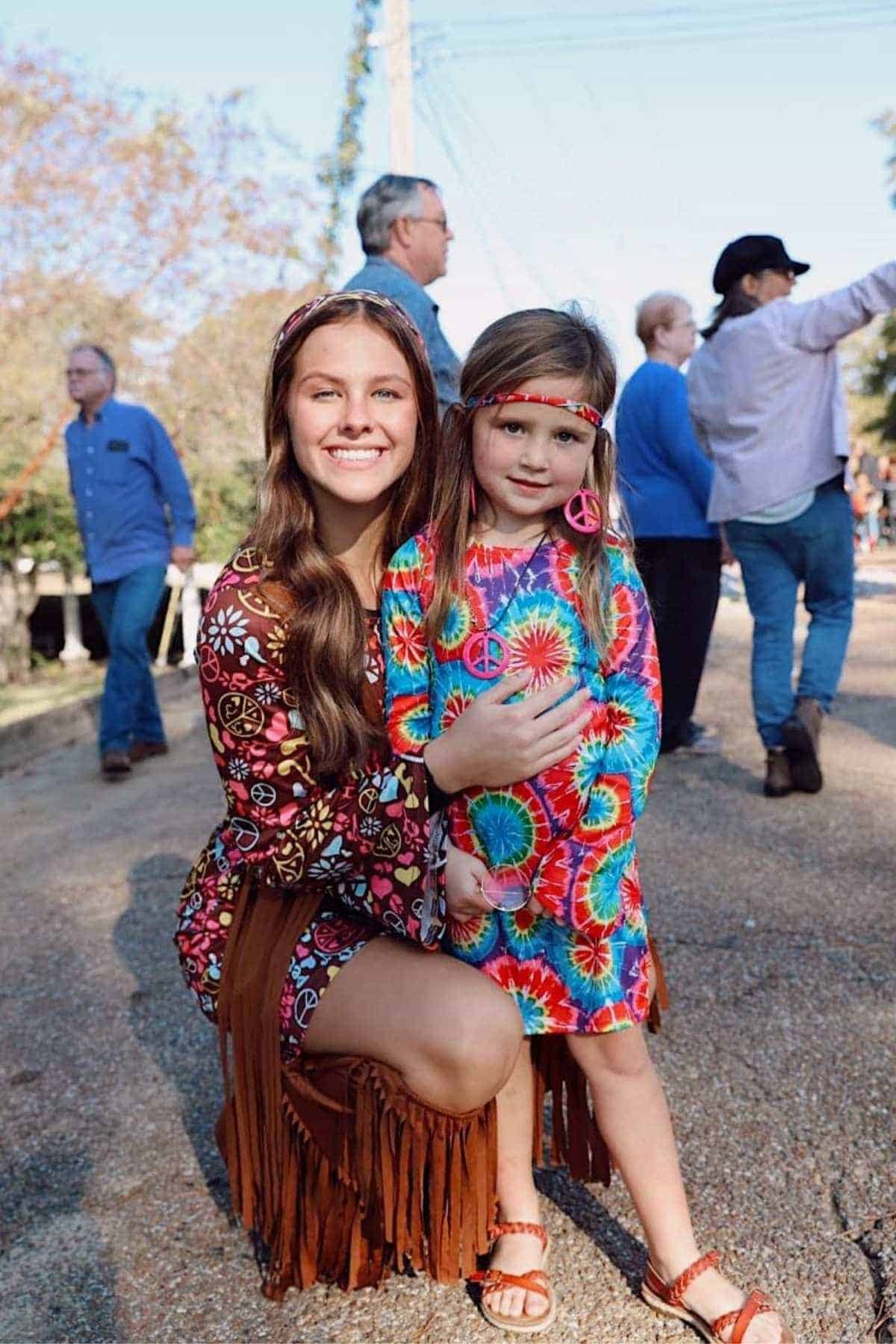 Two girls wearing colorful, retro-style outfits and headbands pose and smile outdoors at a gathering, with people in the background.