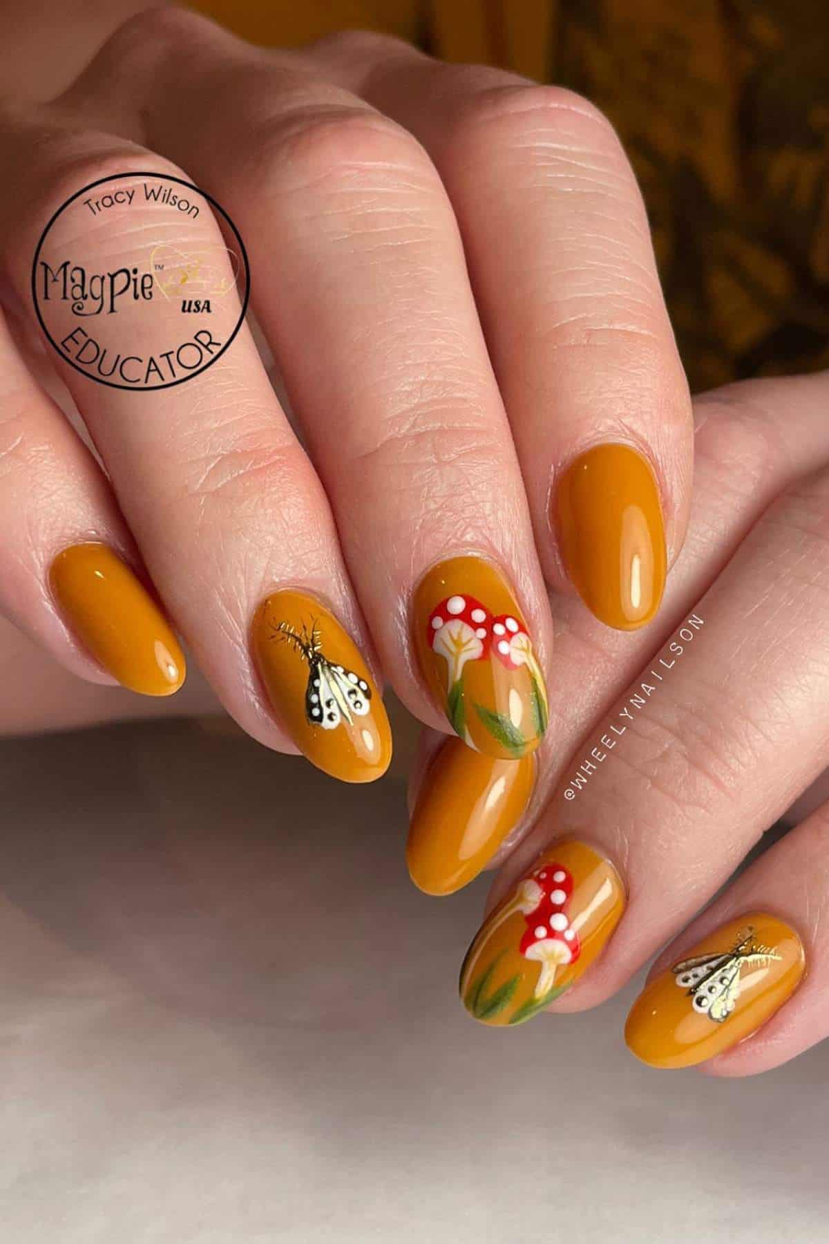 Hand with mustard yellow nails featuring painted designs of butterflies, grass, and red mushrooms with white spots on two accent nails.
