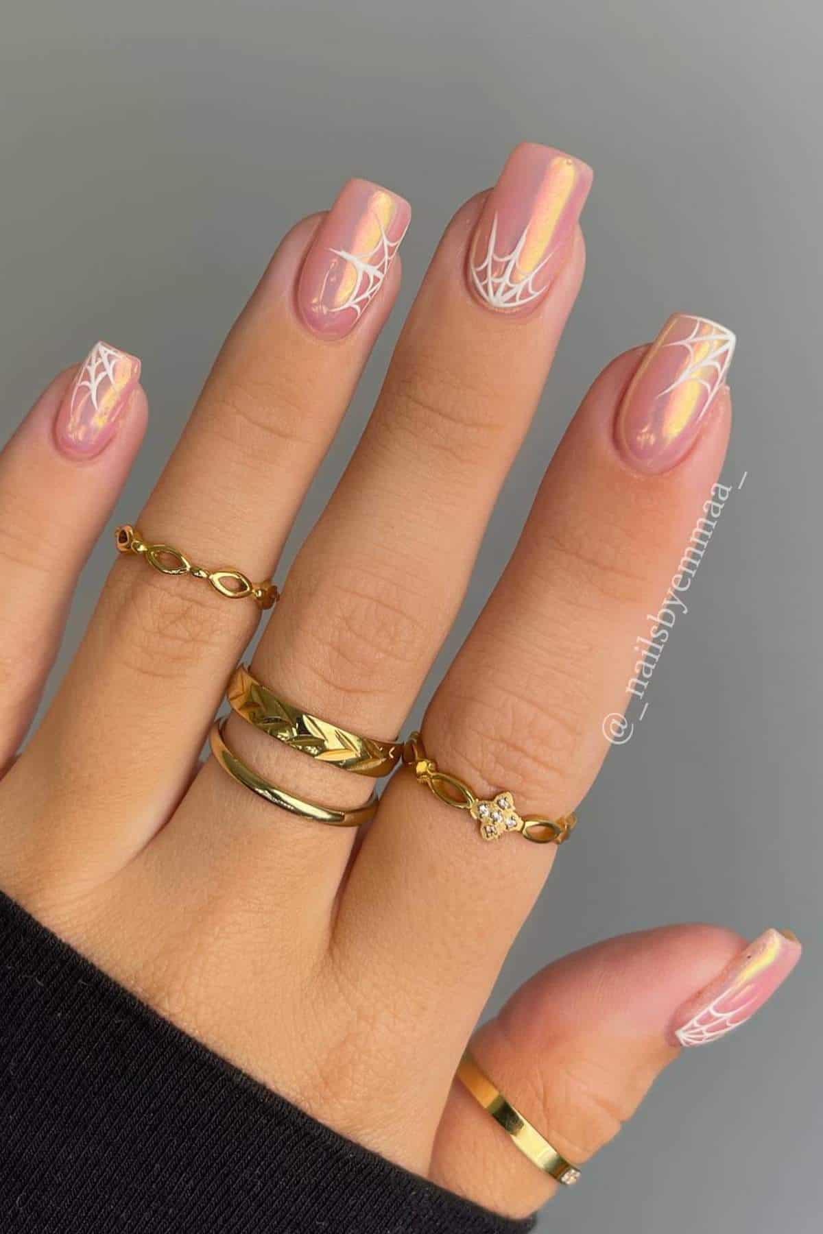 A hand with pink iridescent nails and white spiderweb designs on tips, wearing multiple gold rings, against a neutral background.