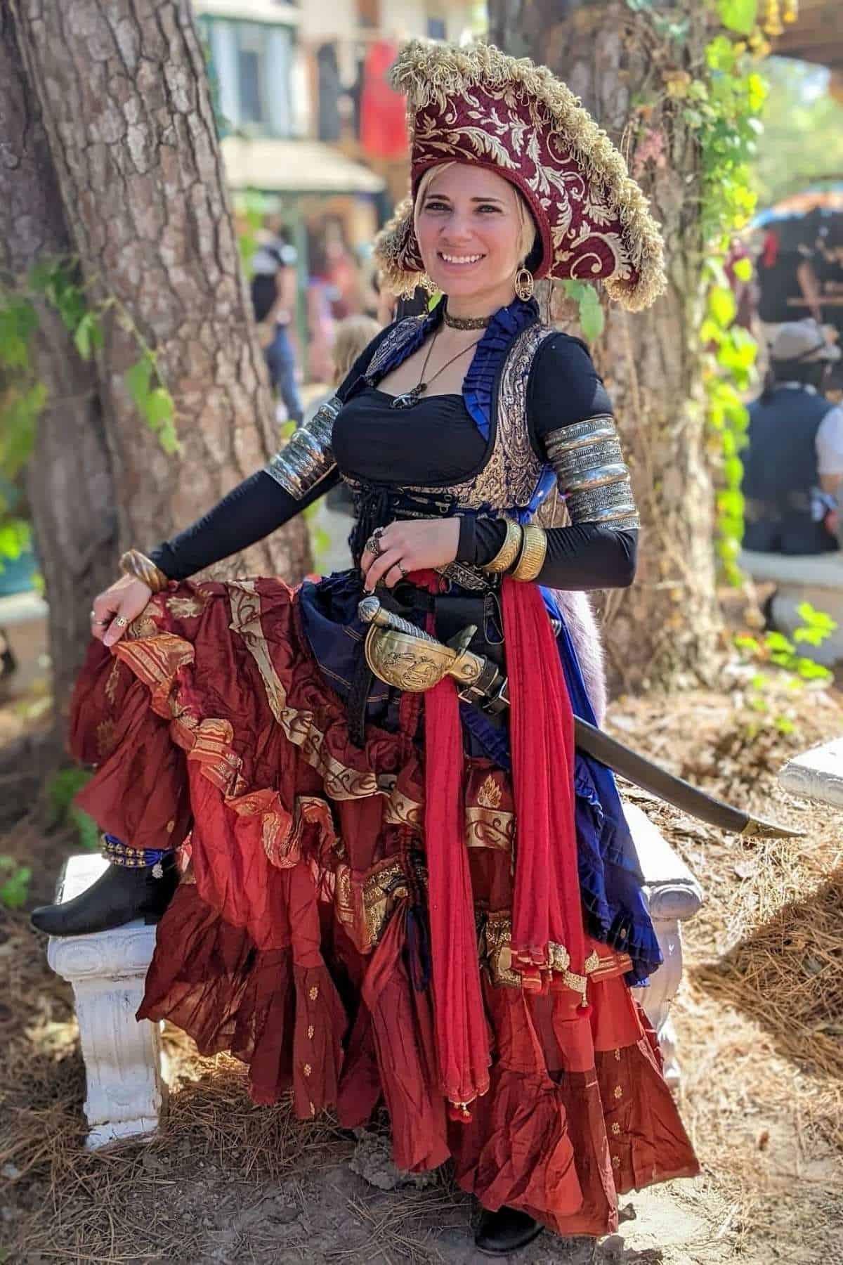 Woman in ornate pirate costume poses outdoors, wearing a feathered hat and layered skirt, with a sword at her waist and a smile on her face.