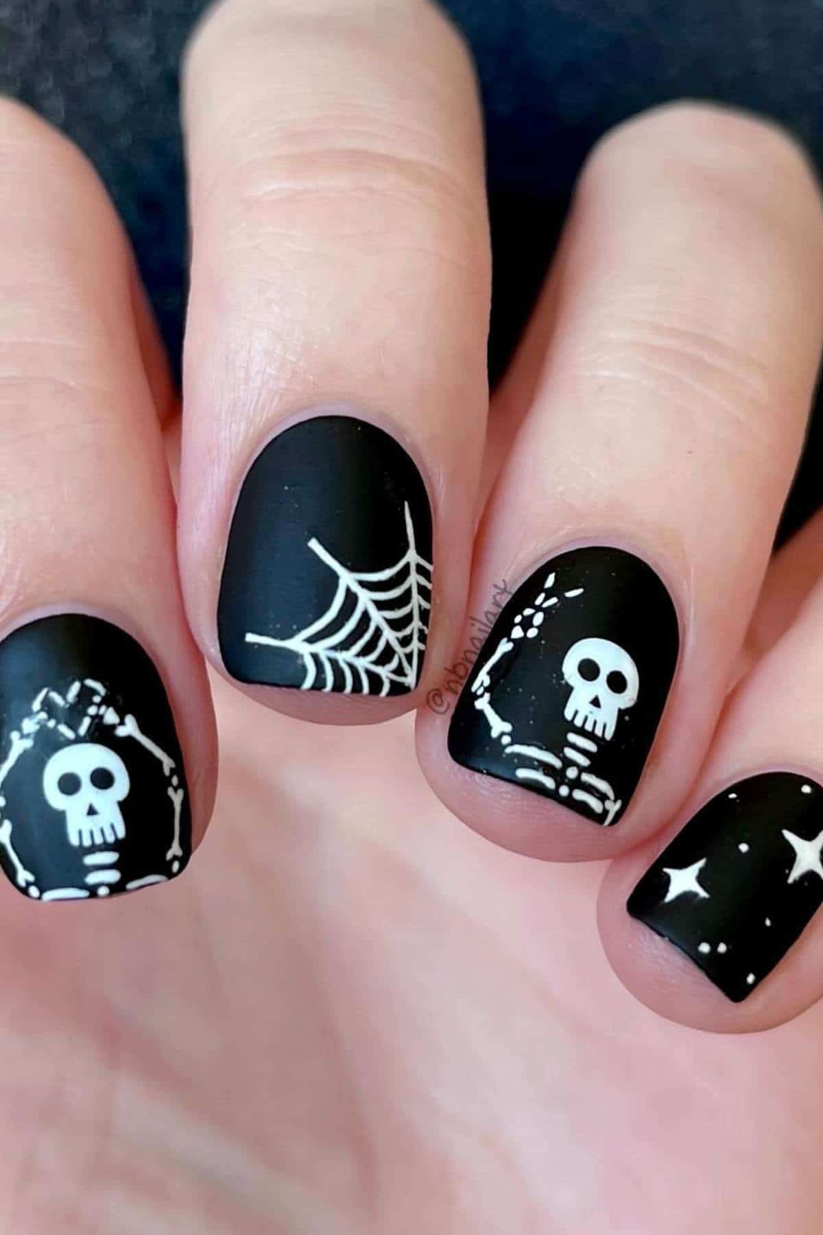 Hand with matte black nail polish featuring white Halloween designs: skulls, webs, bones, and stars.