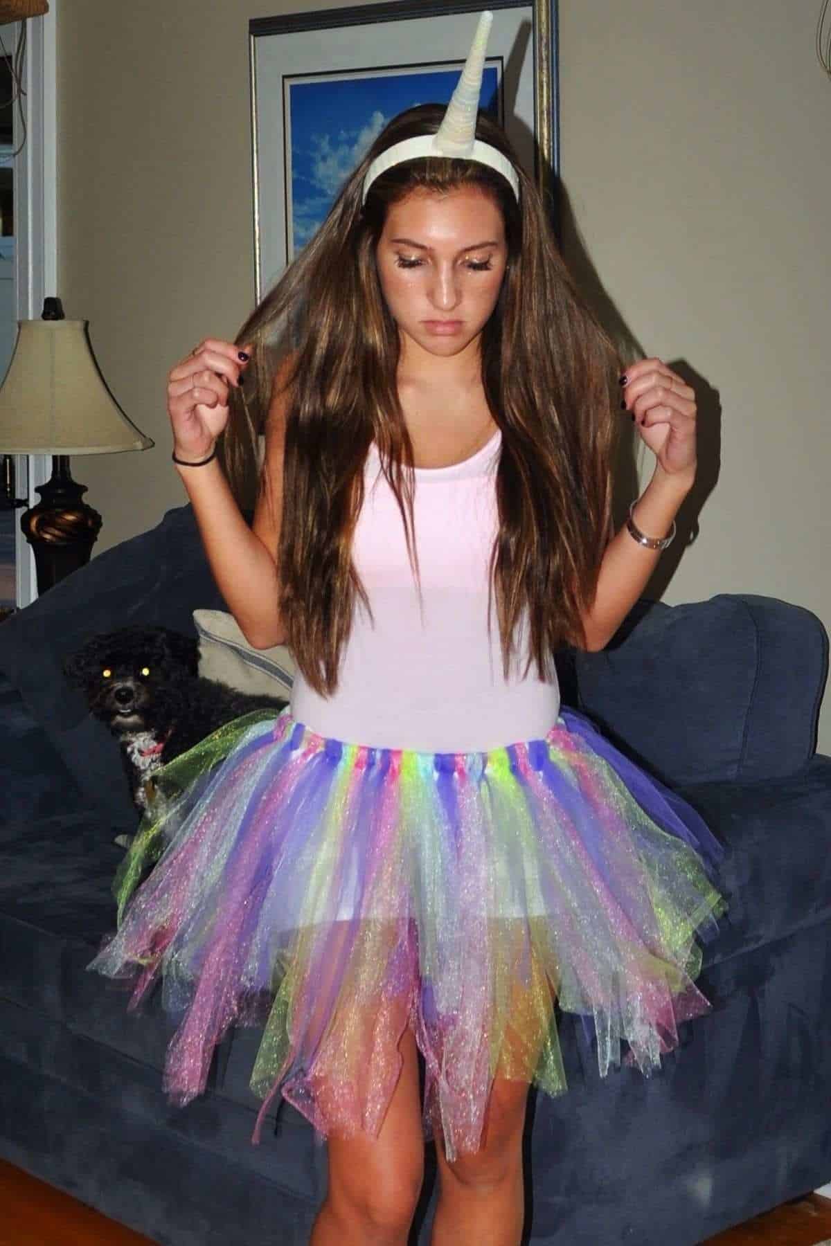 Young woman wearing a unicorn horn headband and a pastel rainbow tutu stands indoors, holding her hair. A small black dog sits on the couch behind her.