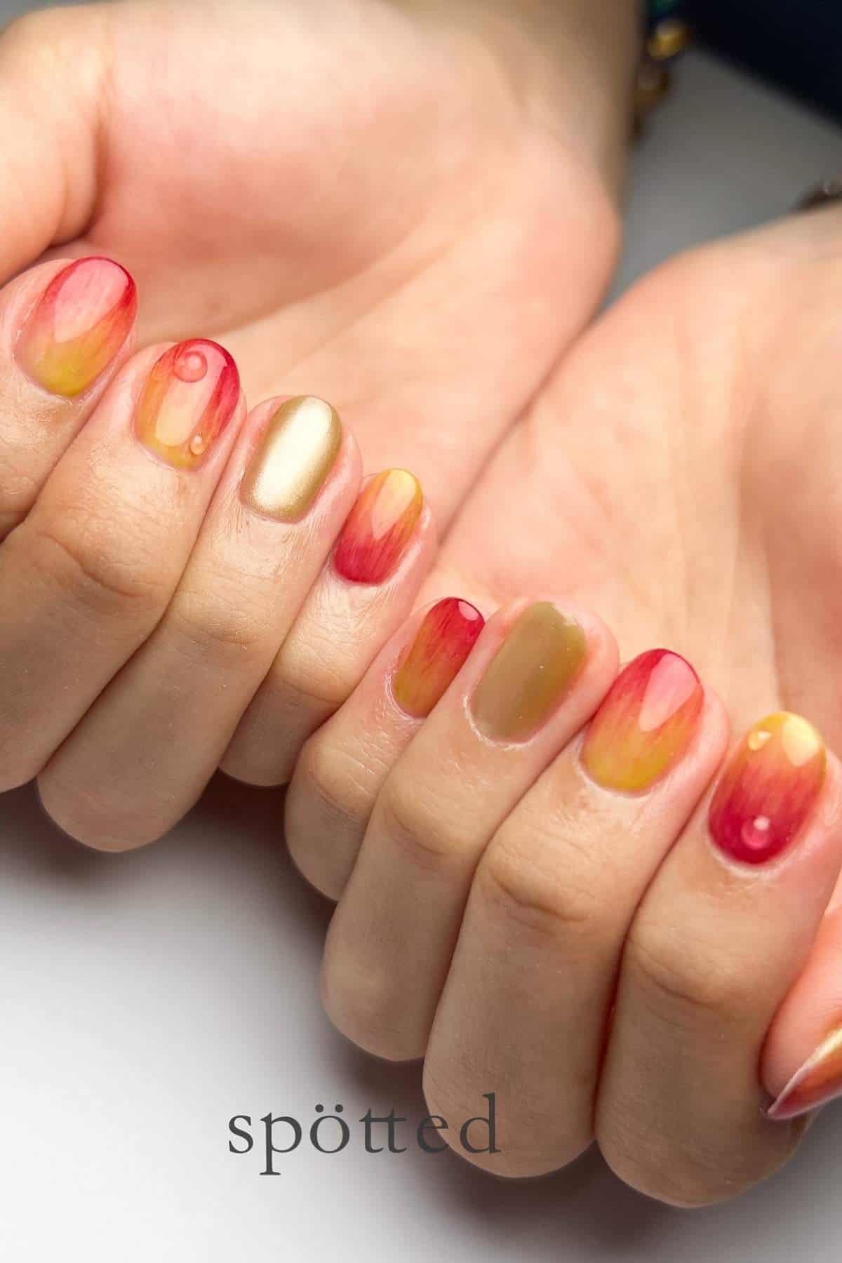 Close-up of hands with a colorful manicure featuring shades of red, yellow, and gold with a glossy finish and abstract, gradient patterns on each nail.
