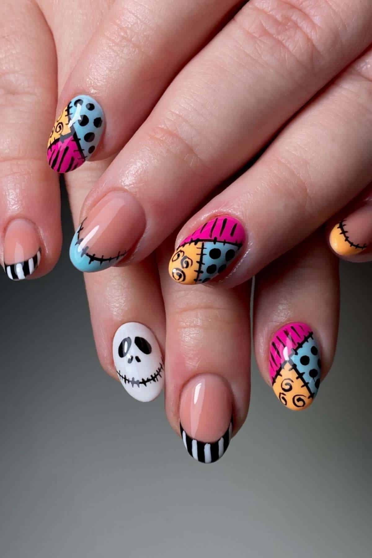 Hands with colorful Halloween-themed nail art featuring patchwork patterns, stripes, and a white nail with a skull face design.
