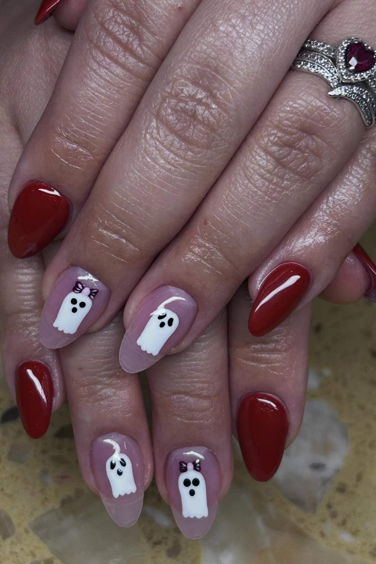 Hands with almond-shaped nails, painted red except for three nude nails featuring white ghost designs with black eyes and mouths, accented by small purple bows.