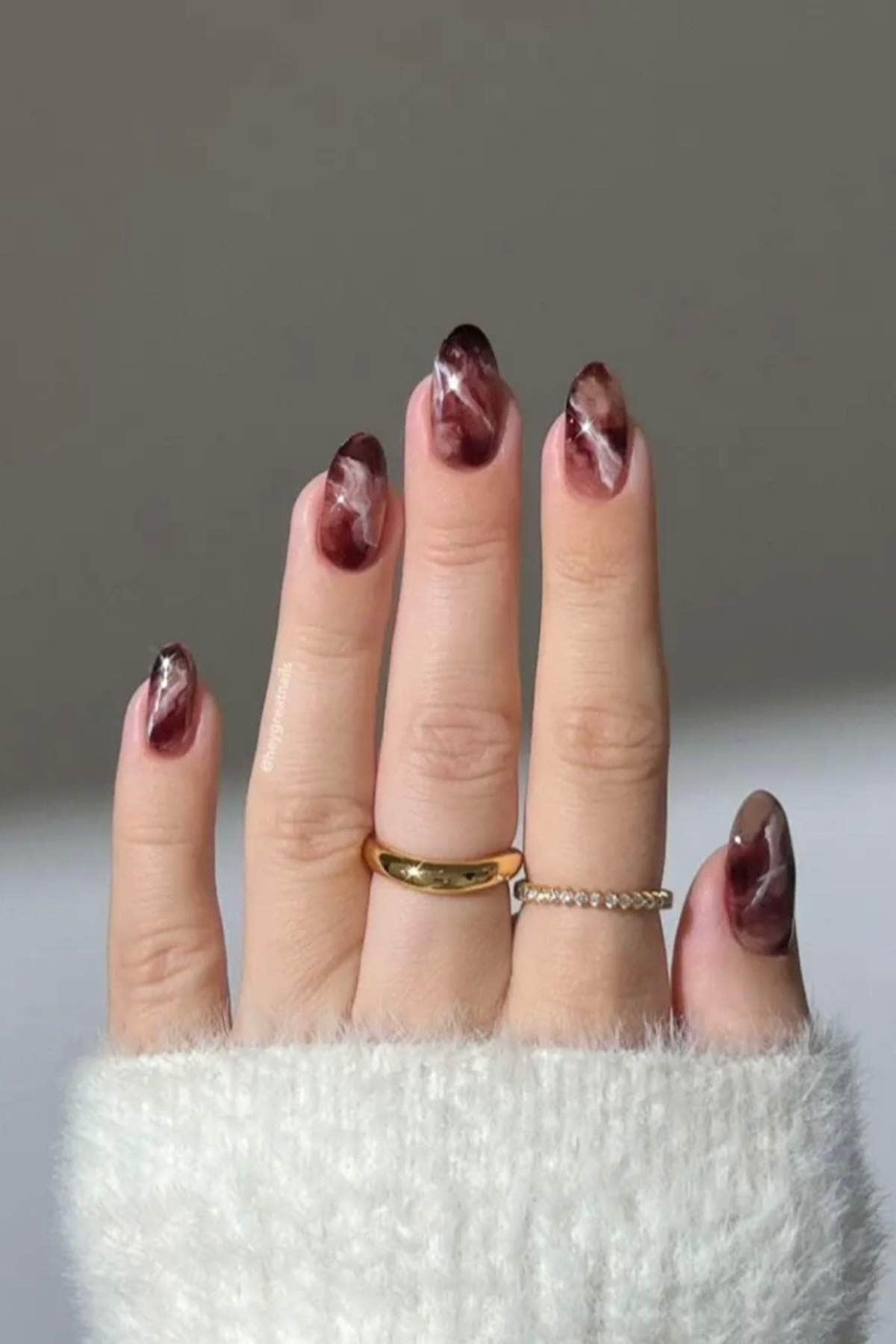 A hand with burgundy and white marble nail art, wearing two gold rings, one plain and one with small stones, against a neutral background.