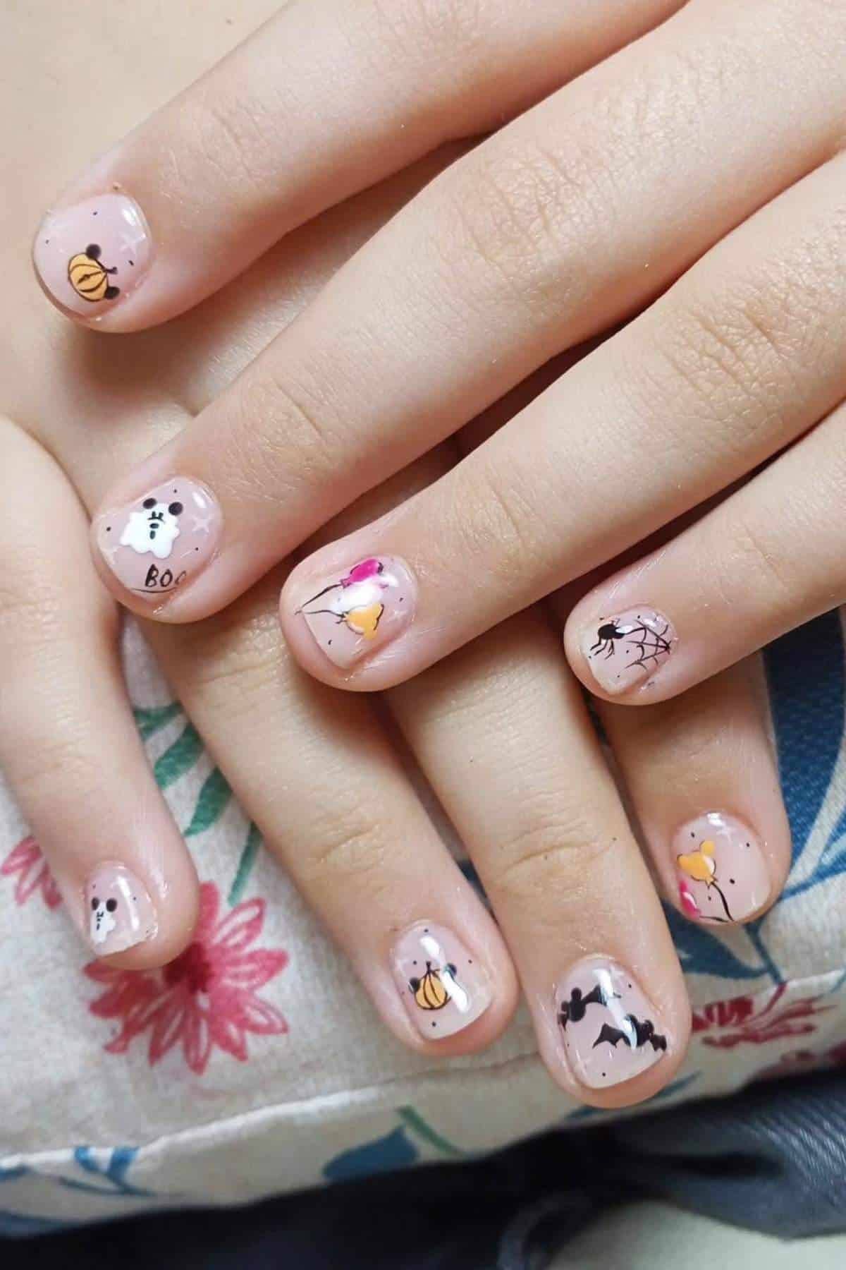 Fingernails with Halloween-themed nail art, including ghosts, pumpkins, bats, a spider, and the word "BOO" on a clear base. Hands rest on a patterned fabric.