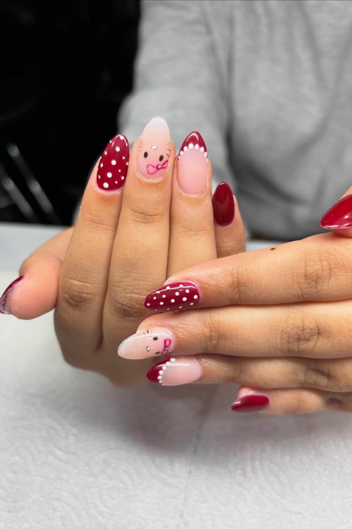 Hands with red and nude manicured nails, featuring white polka dots and cute face designs with pink bows on the ring and index fingernails.