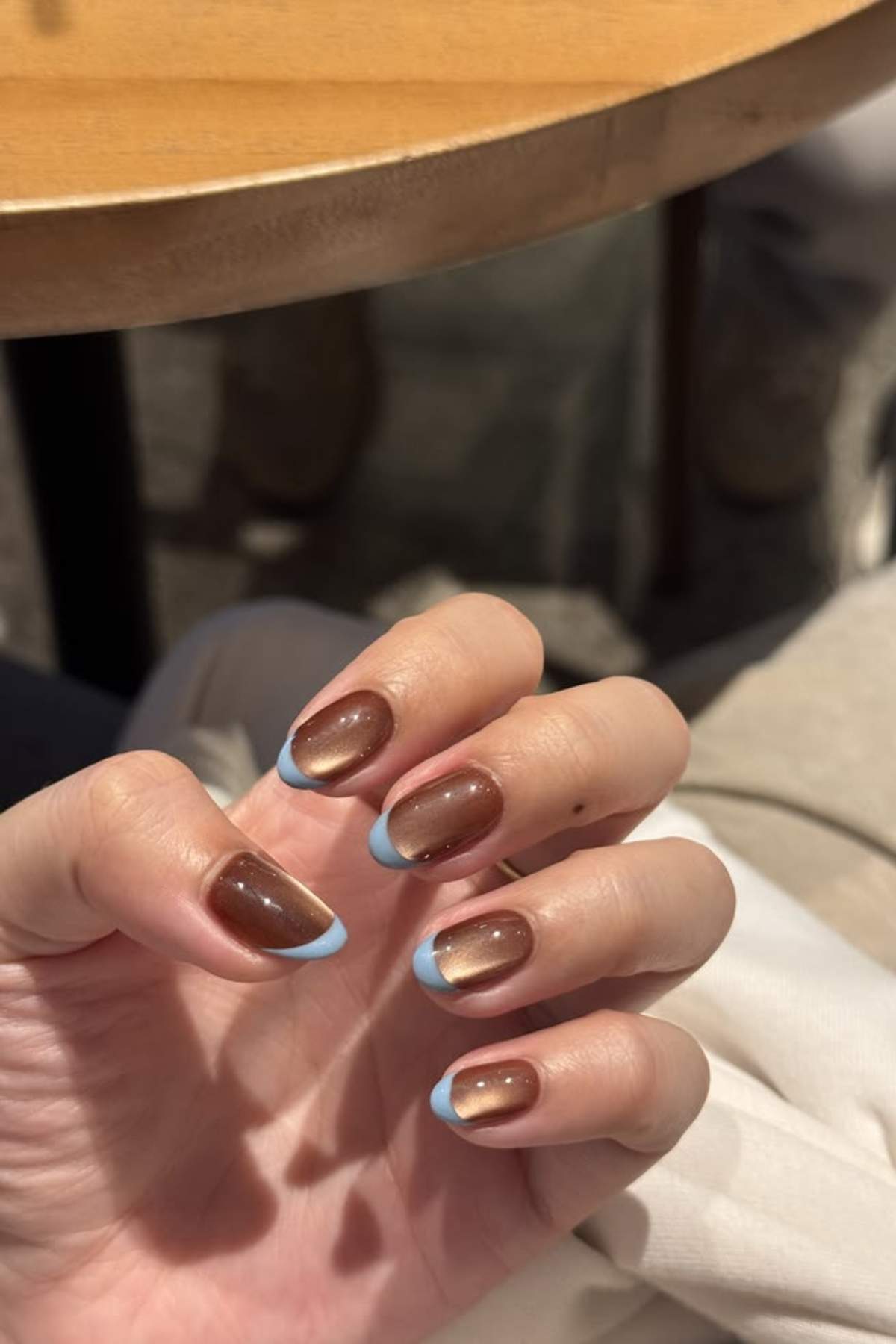 A hand with brown nails featuring light blue French tips is held up under a table.