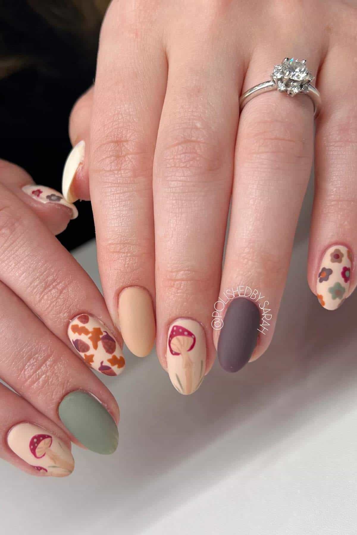A hand with matte, multi-colored nails features various fall-themed designs and a silver ring with a large stone on the ring finger.