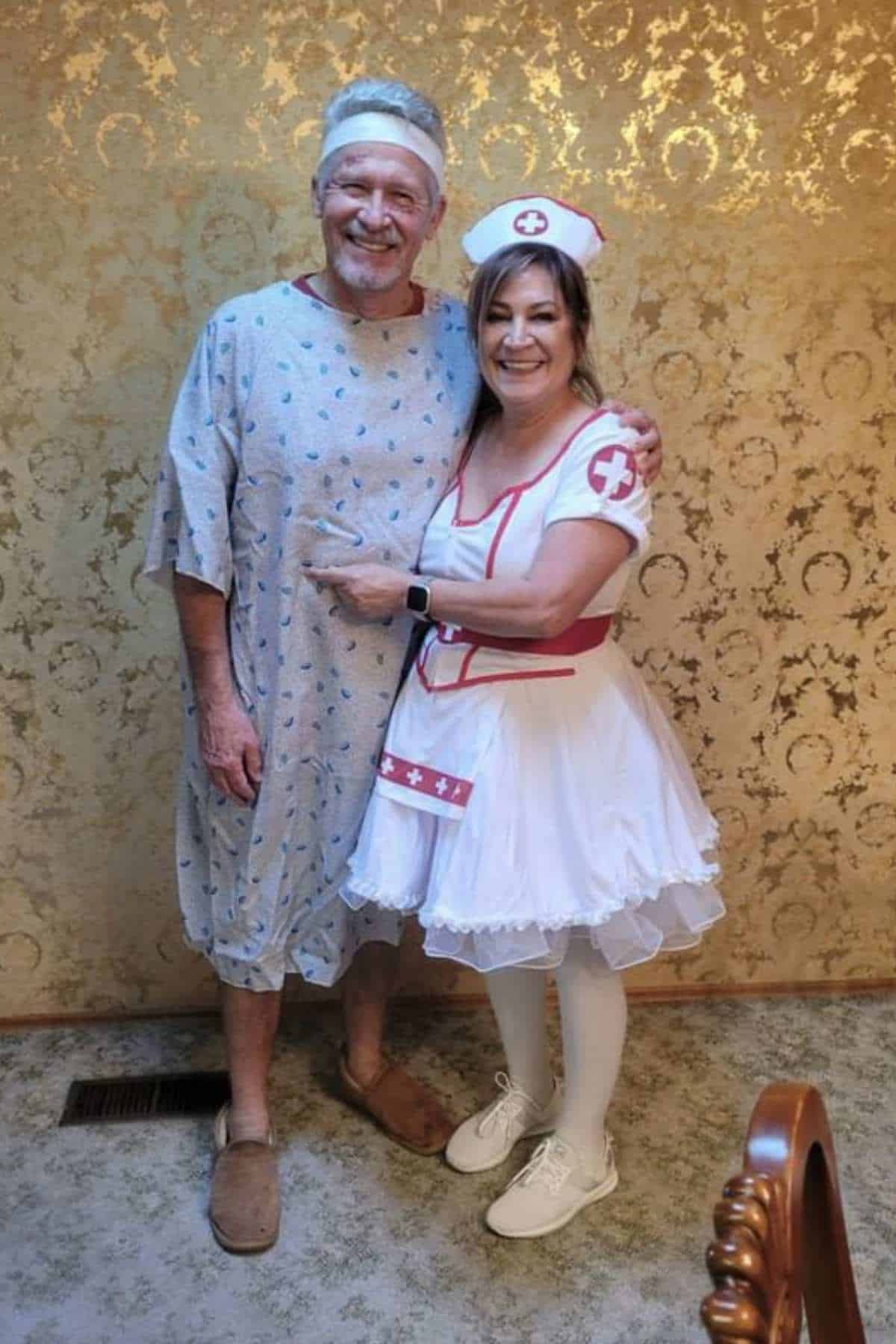 A man in a hospital gown and slippers stands next to a woman in a nurse costume. They are smiling and standing against a patterned gold wall.