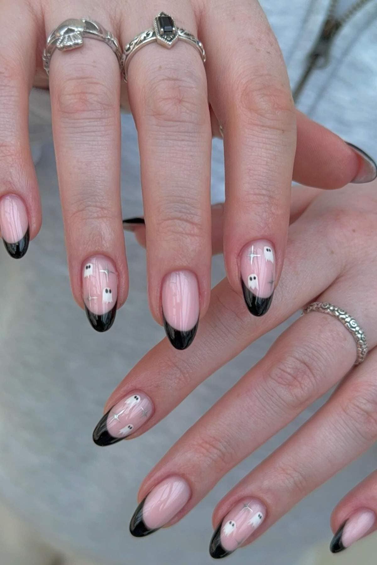 Hands with almond-shaped nails featuring a nude base, black French tips, and small white ghost designs. The person is wearing several silver rings.
