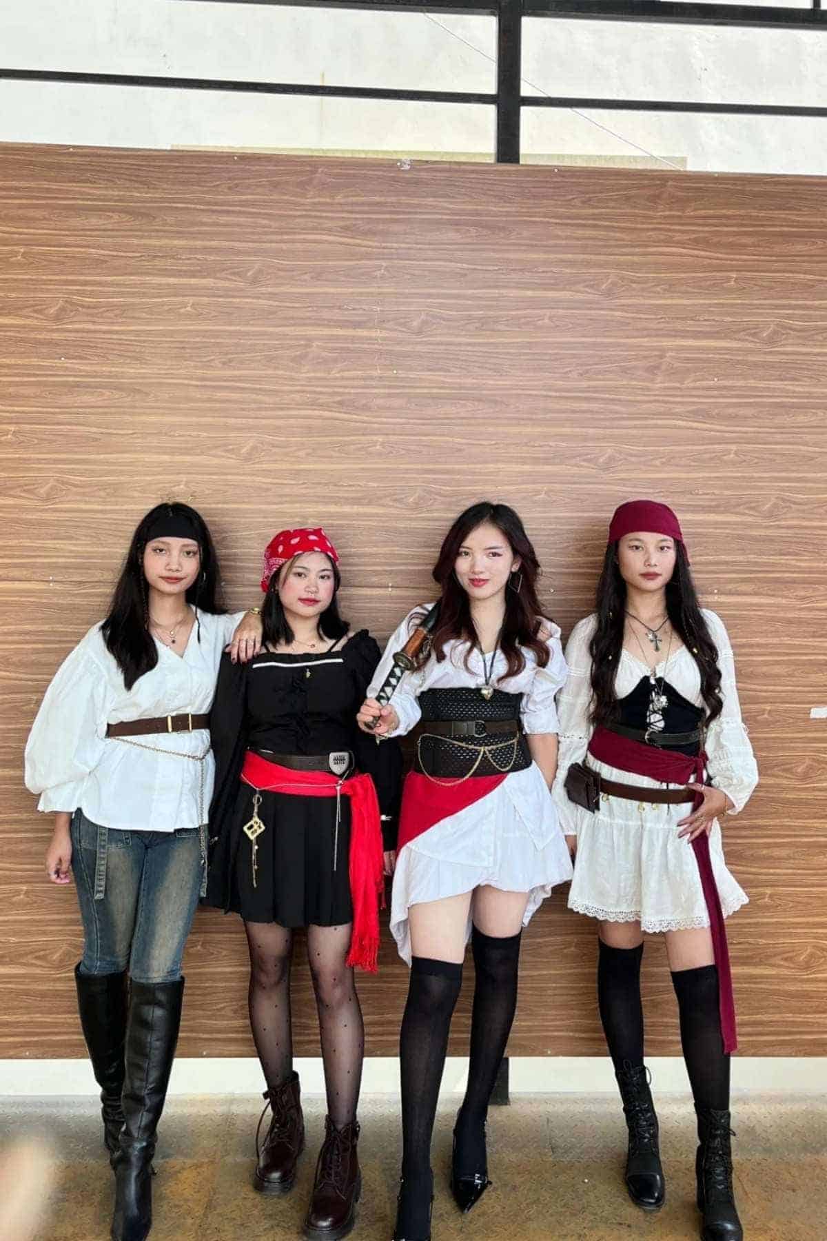 Four women in pirate-themed costumes stand side by side in front of a wooden wall, facing the camera.