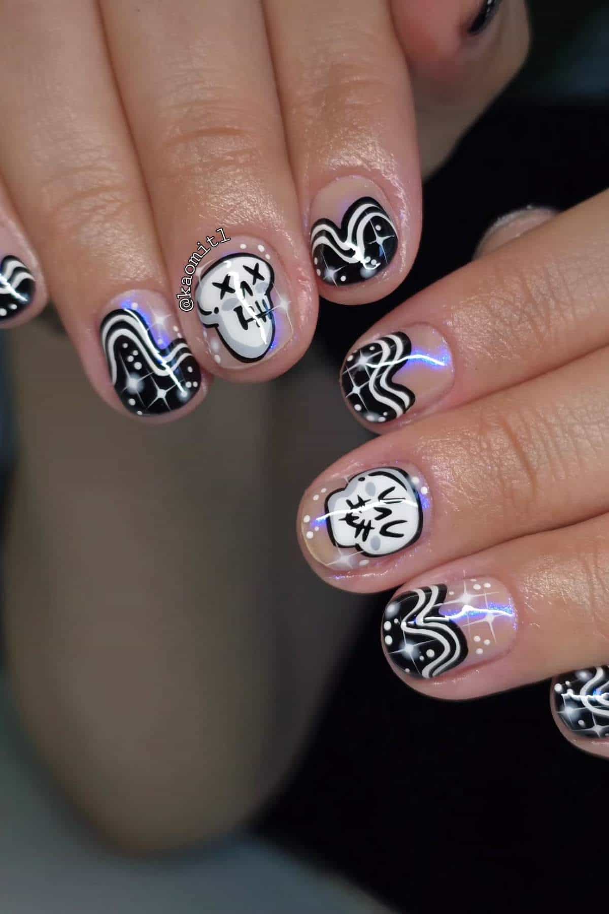 Close-up of hands with black and white nail art featuring wavy lines, dots, a cartoon skull, and a ghost design on a shiny nude base.