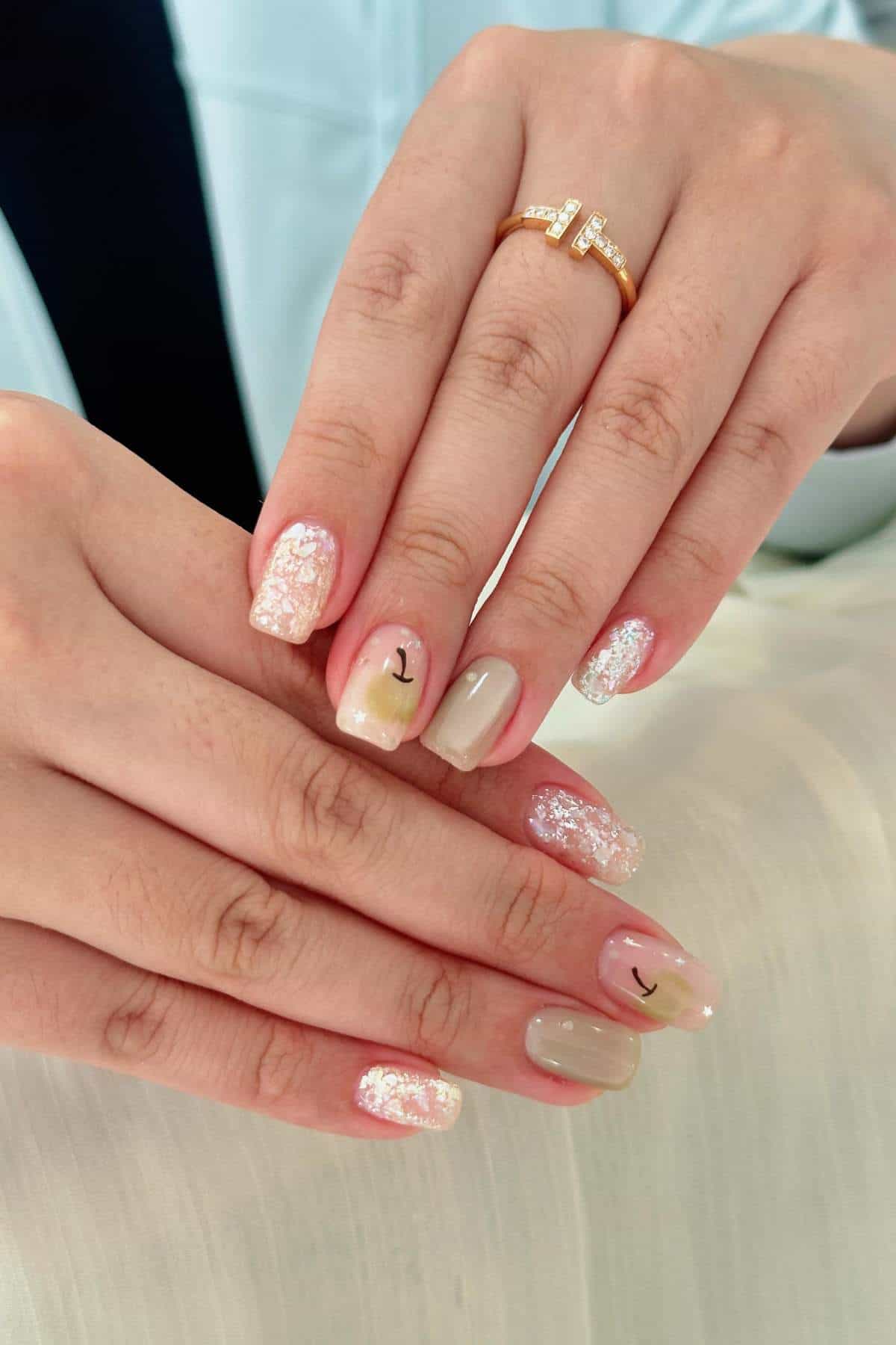 A person with manicured nails featuring nude and pink shades, glitter accents, and minimalistic smiley face designs on two nails, wearing a gold ring with embedded stones.