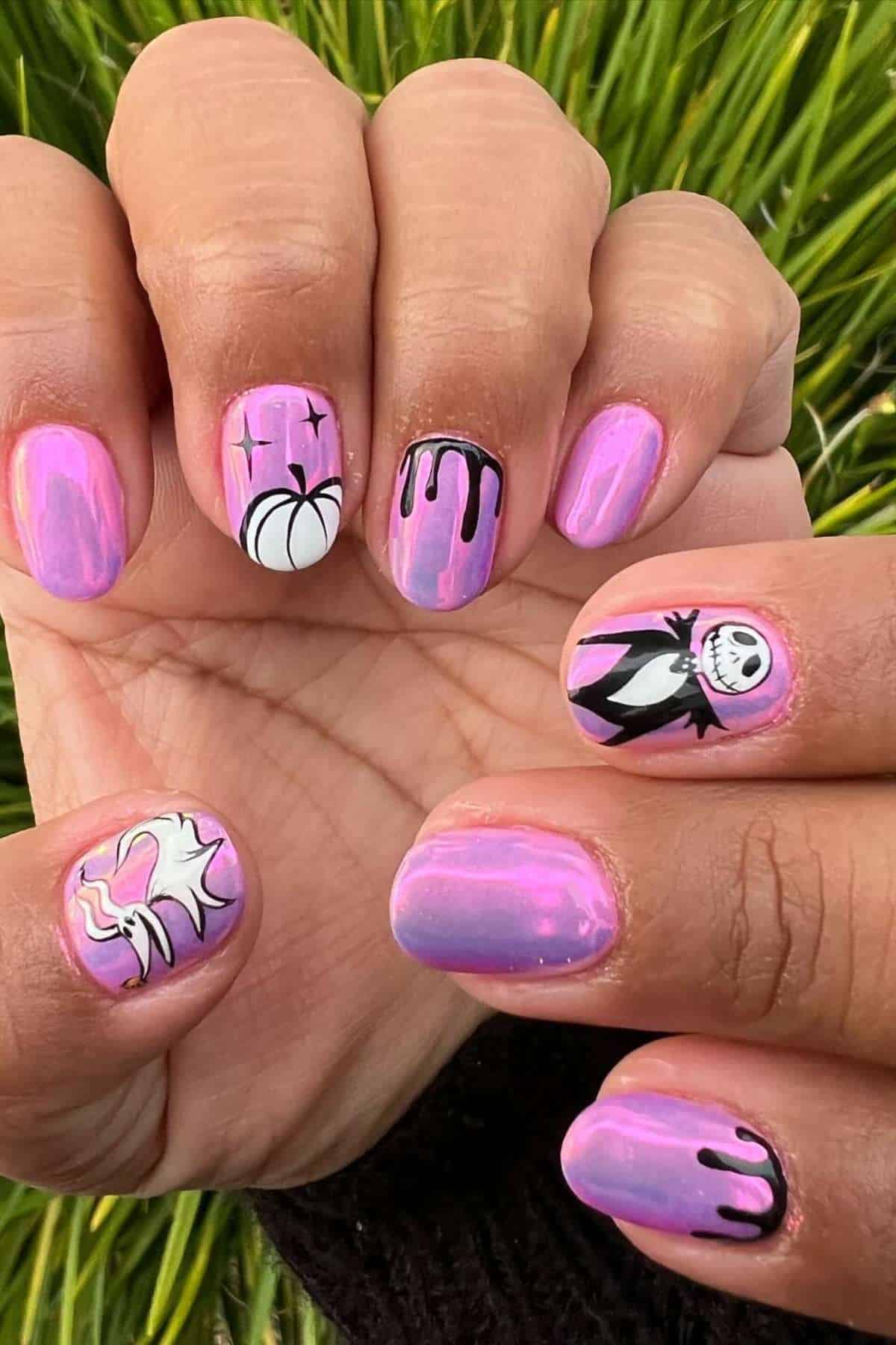 Close-up of hands with iridescent pink nails featuring black drip designs, a white pumpkin, a cartoon skeleton, and a white ghost, set against green grass.