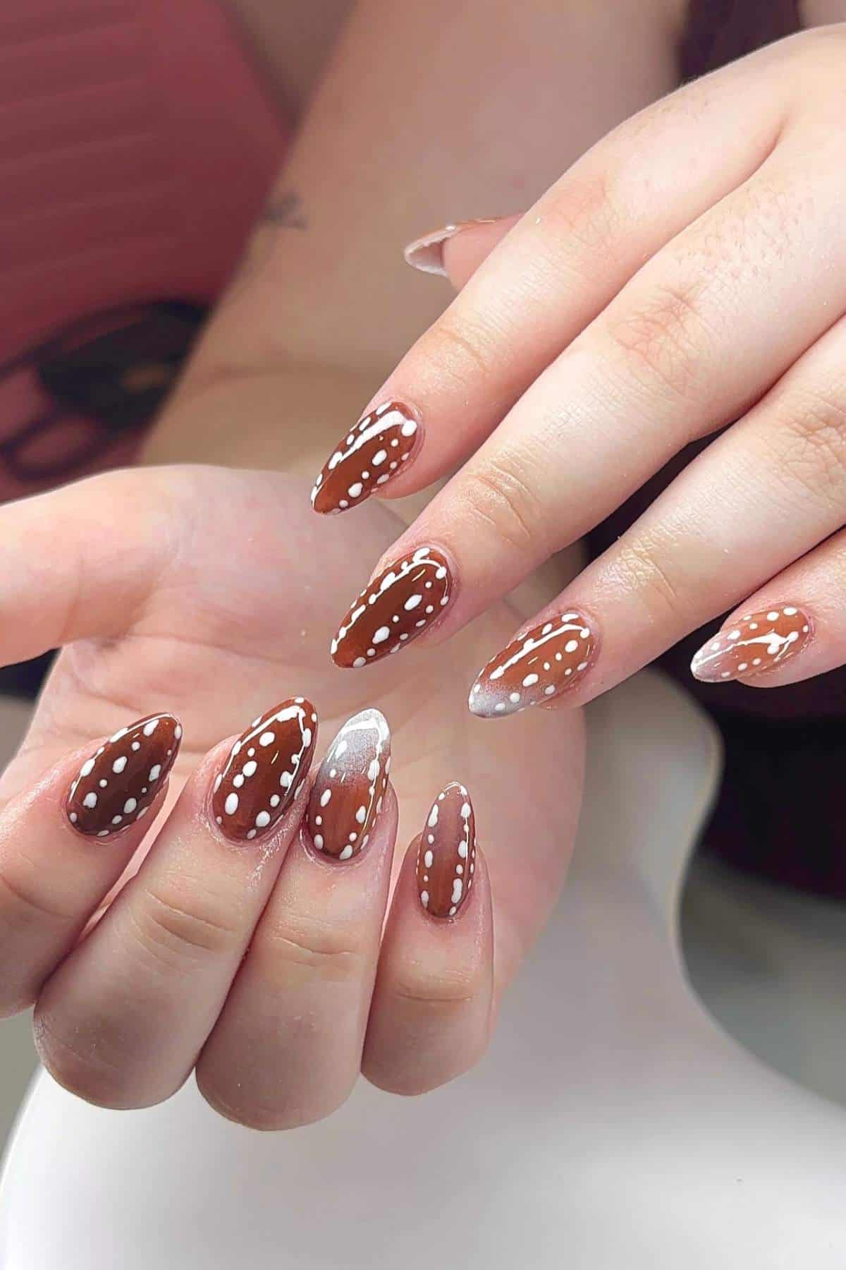 Hands with almond-shaped nails painted brown with white polka dots and one accent nail in white. The background is out of focus.
