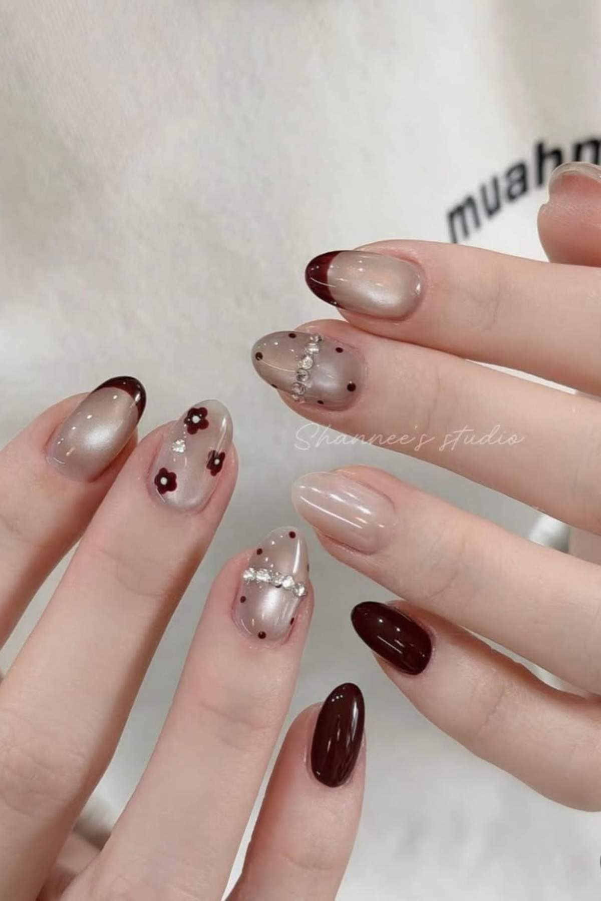 Close-up of hands with almond-shaped nails featuring burgundy tips, floral designs, rhinestones, pearls, and glossy nude polish.