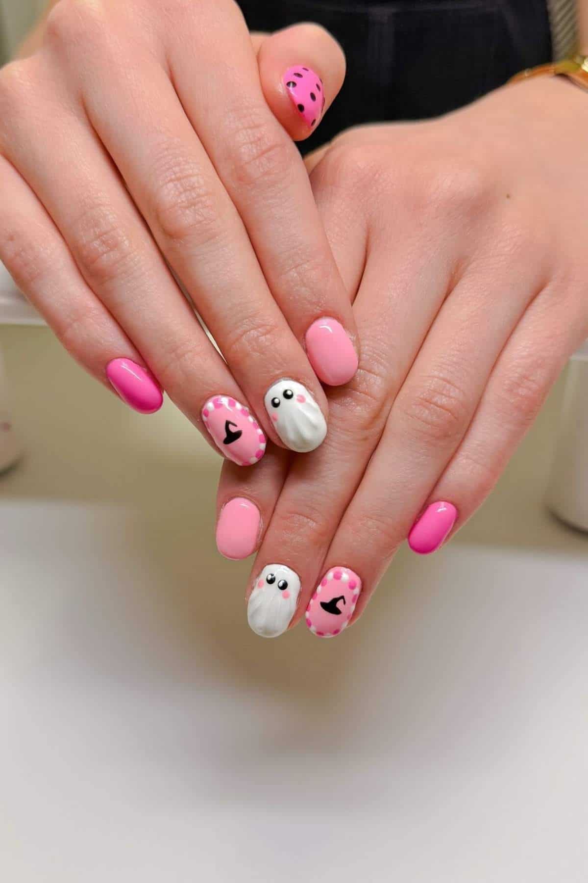 Hands with pink and white nail art; some nails feature black heart shapes and others depict white ghosts with eyes on a pink background.
