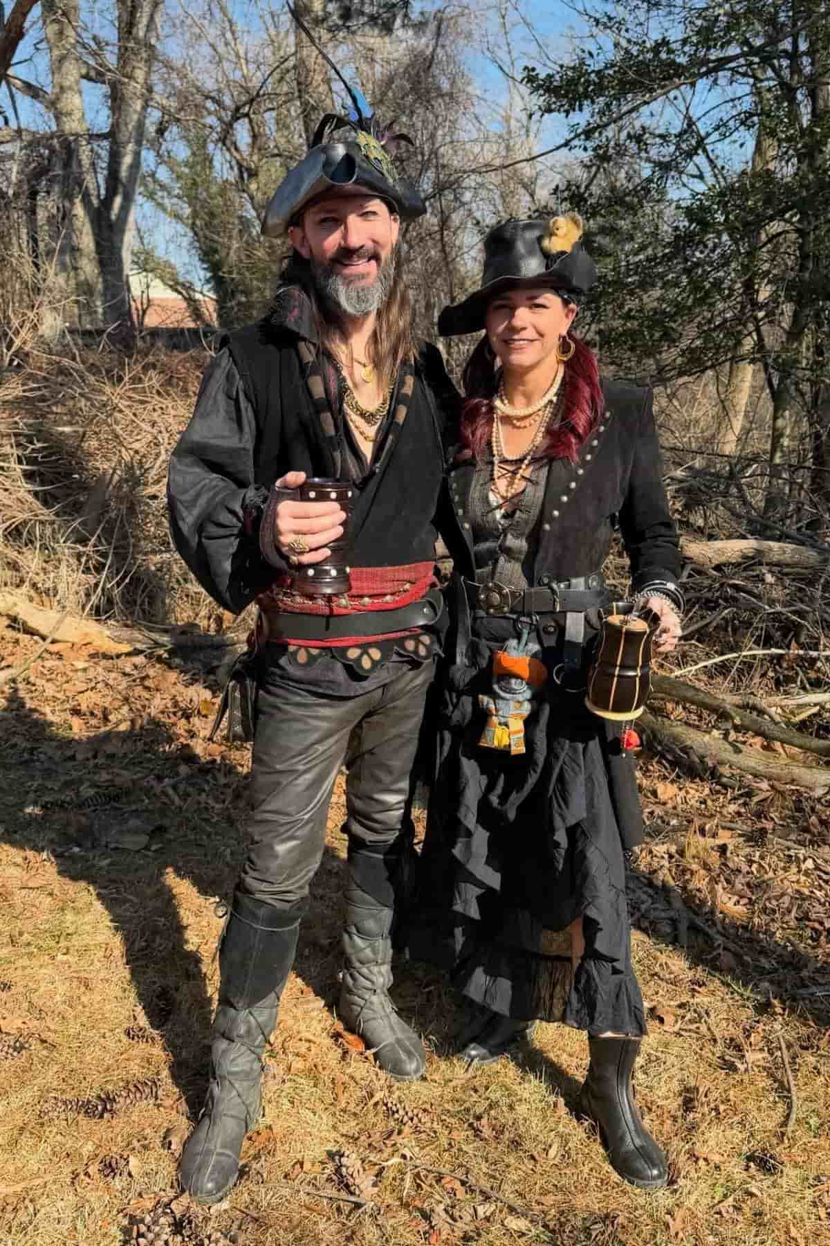 Two adults dressed in pirate-themed costumes stand outdoors on grass, smiling at the camera. They wear black clothing, hats, boots, and carry various accessories. Trees and branches fill the background.