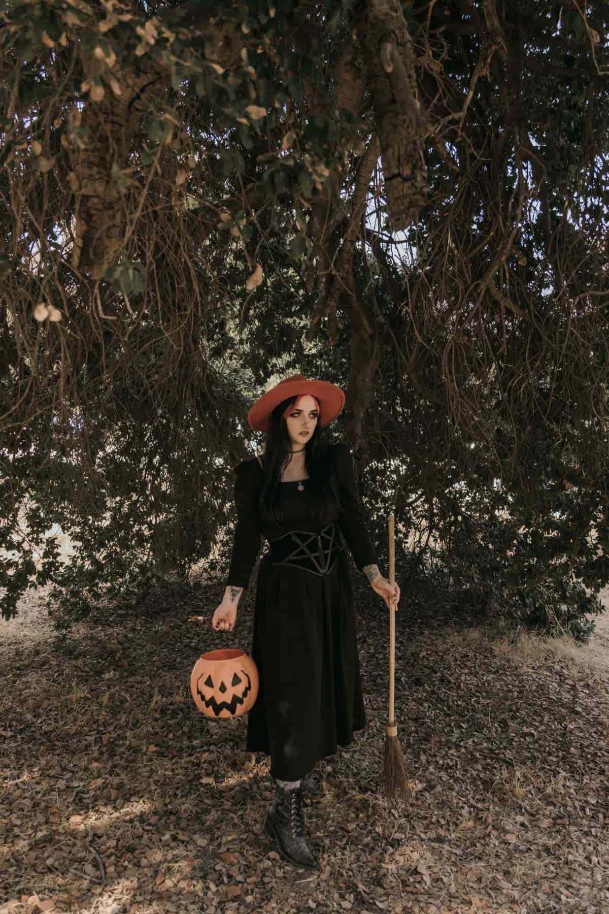 Person in a black dress and red hat holds a broom and a jack-o’-lantern bucket, standing under a large tree with low-hanging branches.