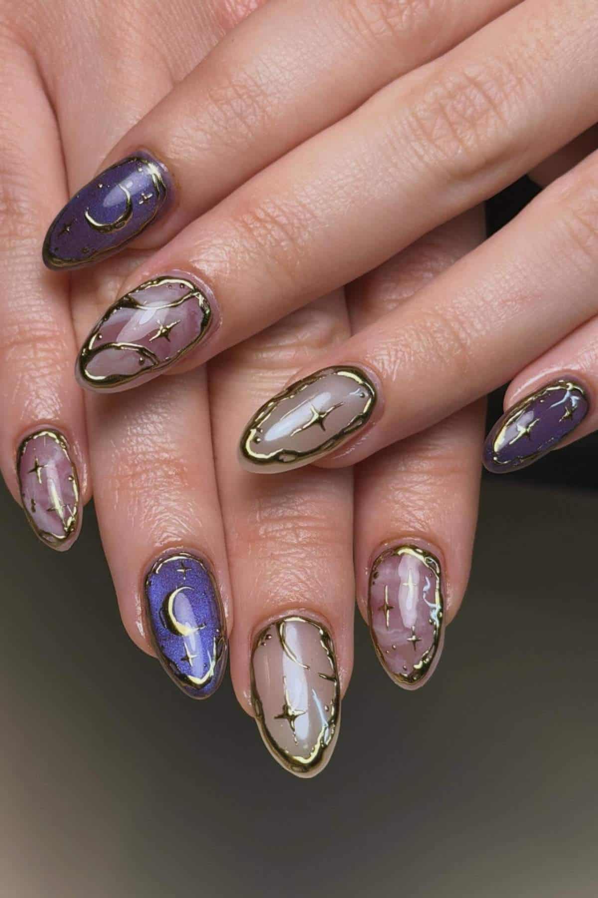 Hands with almond-shaped nails featuring purple and nude marble designs, gold outlines, and celestial motifs like moons and stars.