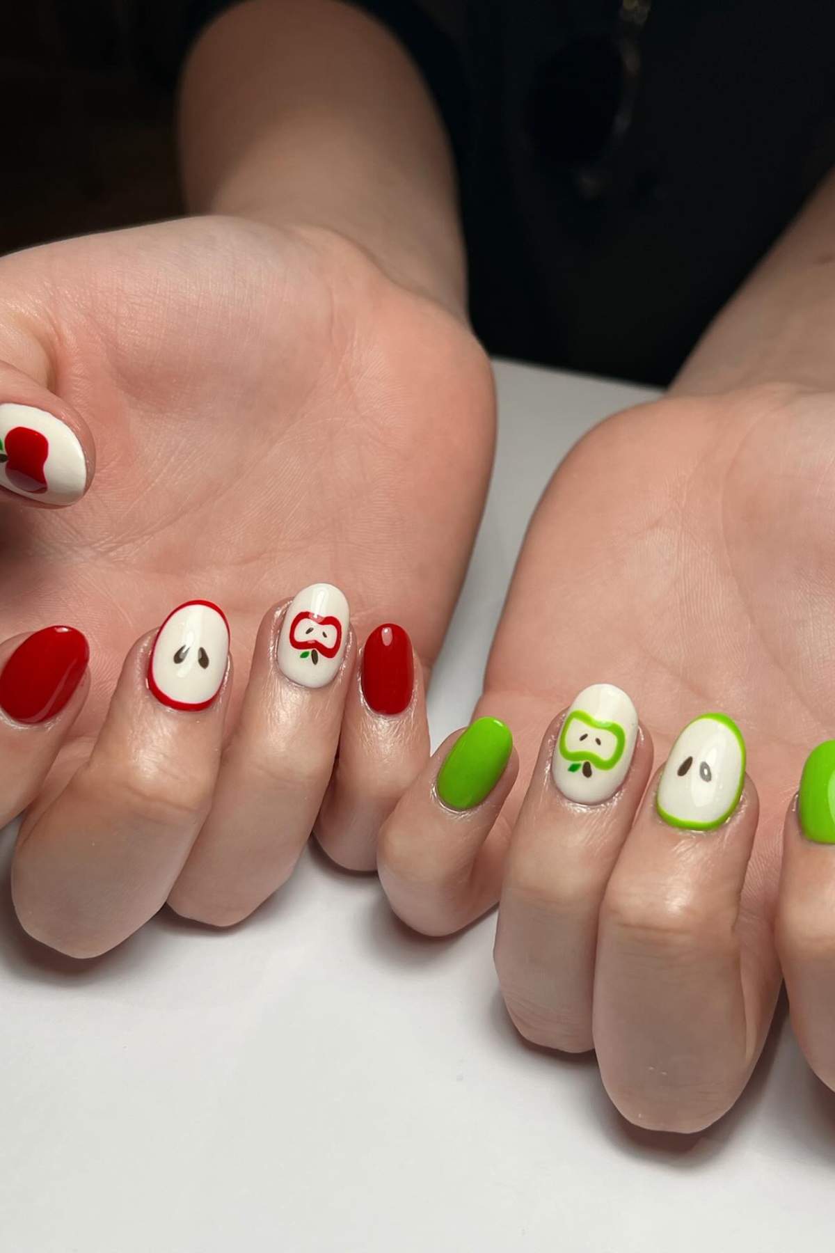 Hands with manicured nails featuring apple-themed nail art in red, green, and white, with some nails painted as apple halves and seeds.