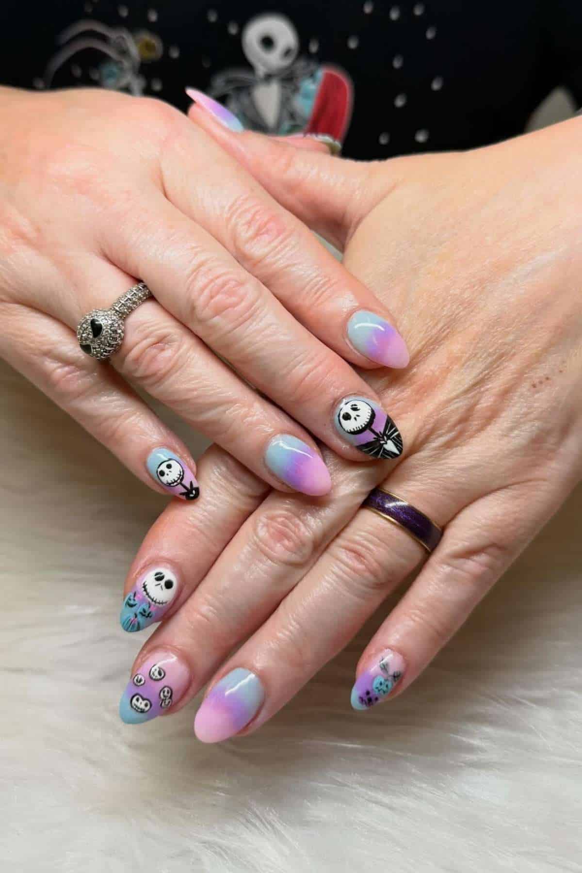 Hands with pastel ombré and "Nightmare Before Christmas" inspired nail art, featuring Jack Skellington faces and themed designs. The person is wearing skull rings.