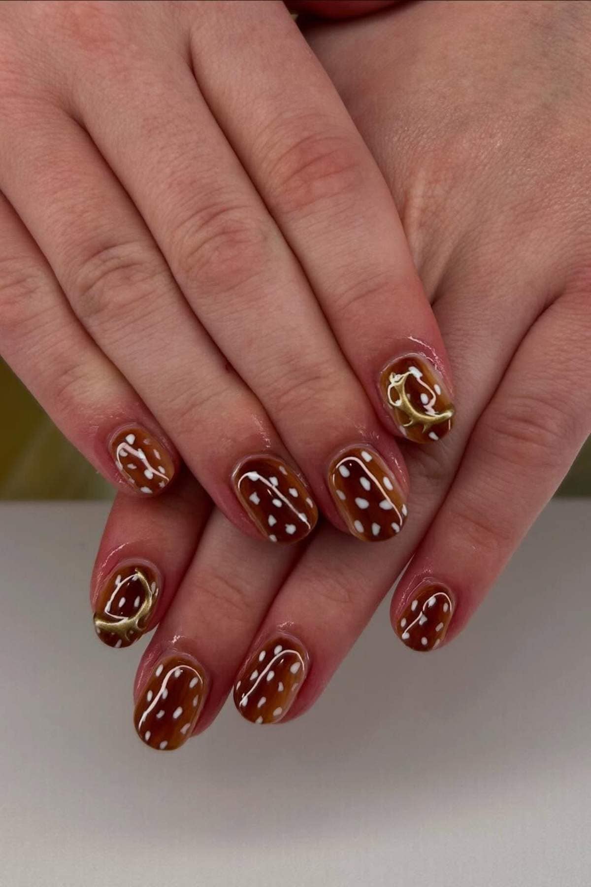 Hands with brown nail polish featuring white polka dots and wavy white and yellow line designs on short, oval-shaped nails.