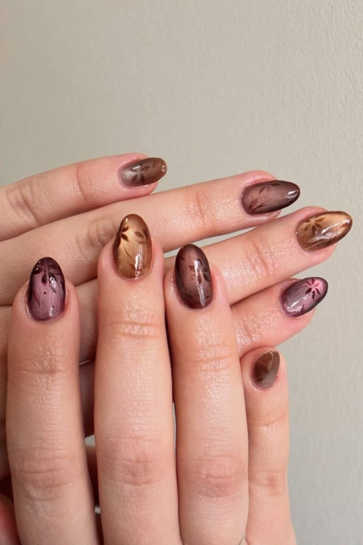 Hands with oval-shaped nails painted in shades of brown, featuring abstract floral and leaf designs on each nail, held against a plain beige background.